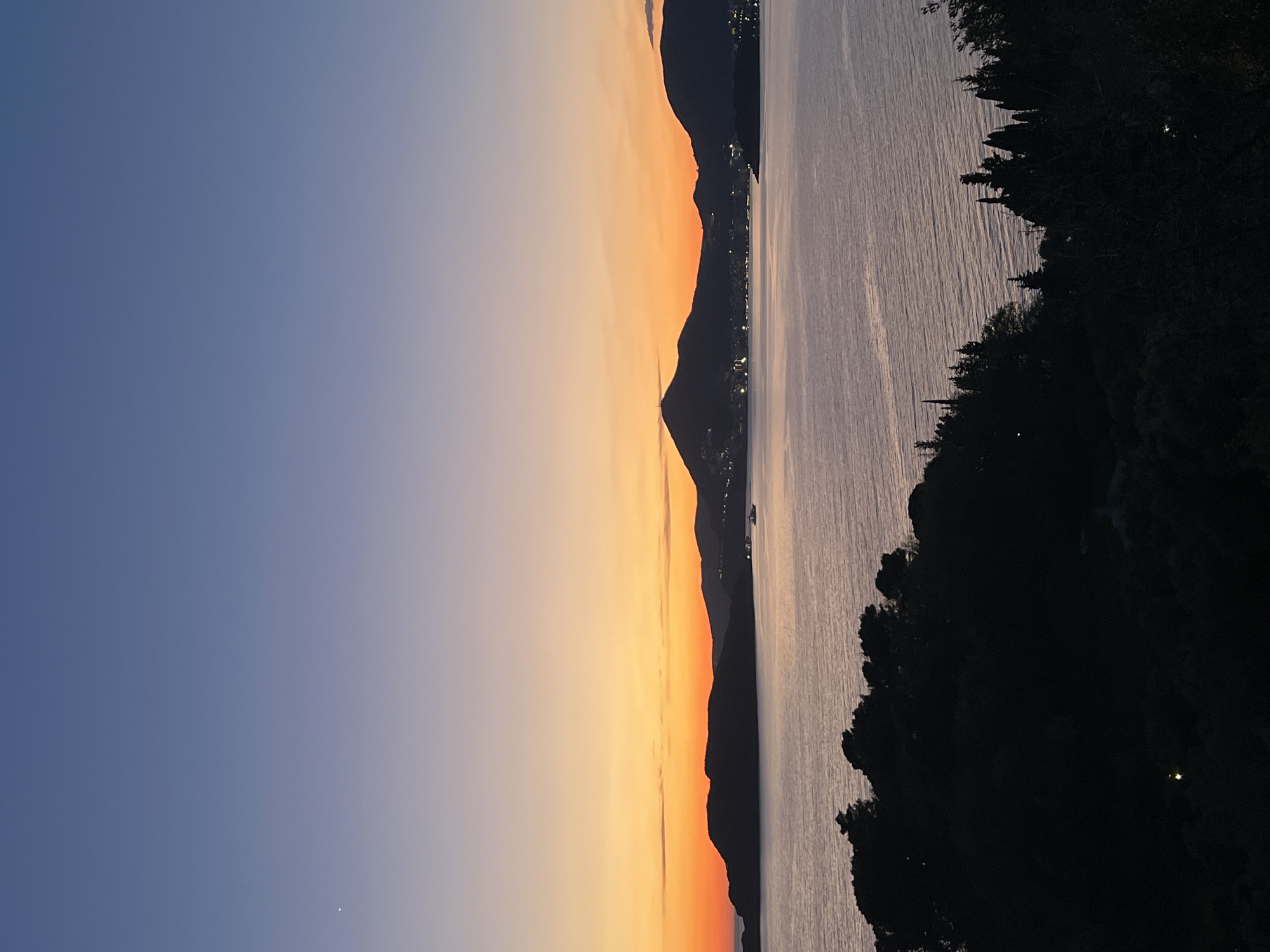 The image captures a coastal view at dusk, likely in or near Sveti Stefan, Montenegro, given the landscape. The left of the frame showcases a gradient sky transitioning from deep blue to orange near the horizon, suggesting either sunrise or sunset. A mountain range is silhouetted against the orange glow. To the right, the sea reflects the fading light. A dark, shadowy mass representing vegetation or land juts into the right side of the image, obscuring some of the water. Lights are visible in the distance on the mountains indicating a city.