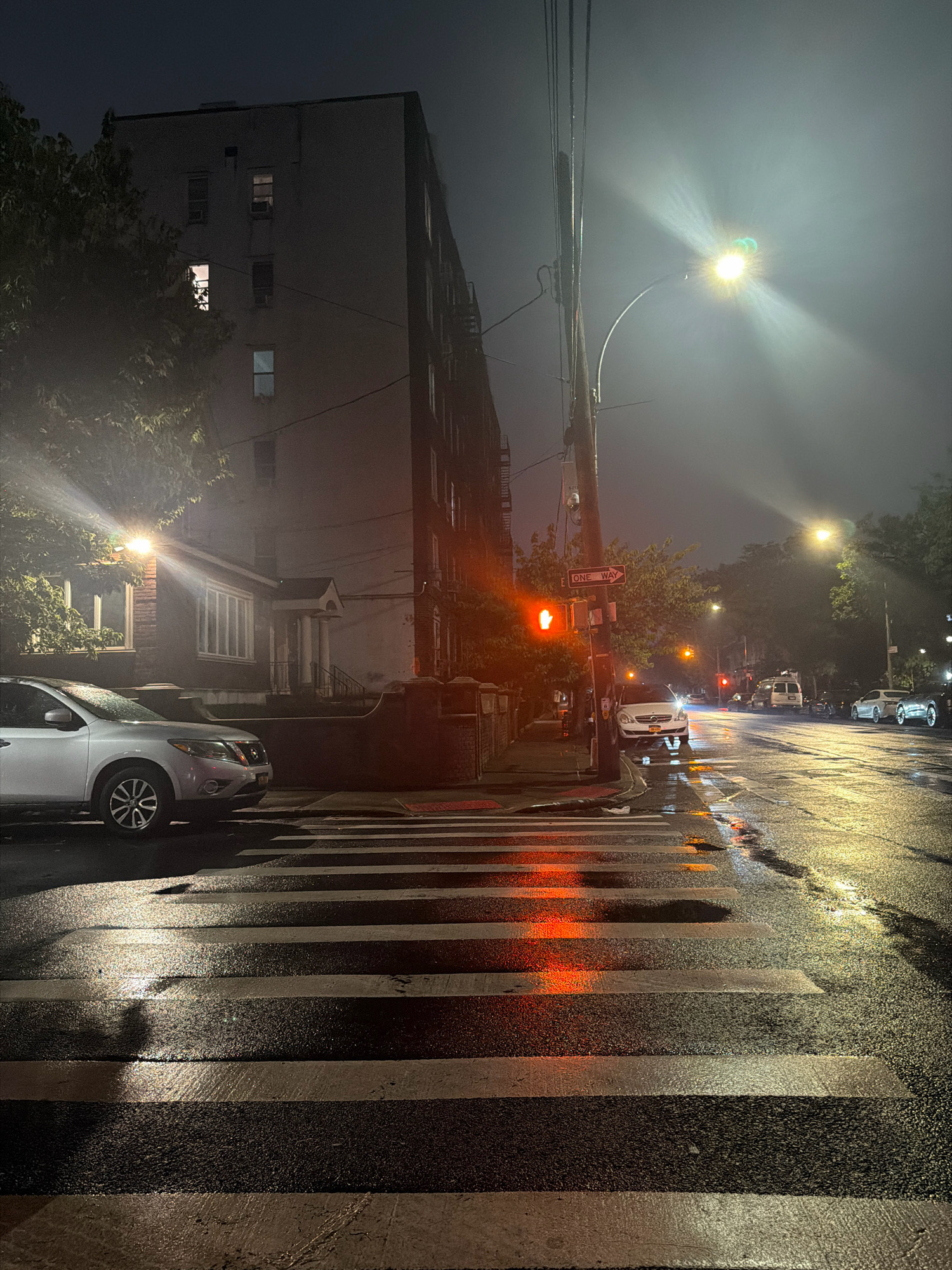 This is a photograph of a street scene at night in New York City, United States of America. The image depicts a wet street with a crosswalk in the foreground, reflecting the lights of passing vehicles and streetlights. A silver SUV is parked to the left of the crosswalk, and another white car is parked further down the street. A building with several illuminated windows is visible on the left side of the image. Streetlights and traffic signals are present, with one traffic light showing a red hand, indicating "Don't Walk." The street appears to be in a residential or mixed-use area, with trees and a building lining the sidewalk. The overall atmosphere is moody and atmospheric due to the nighttime setting and wet conditions.