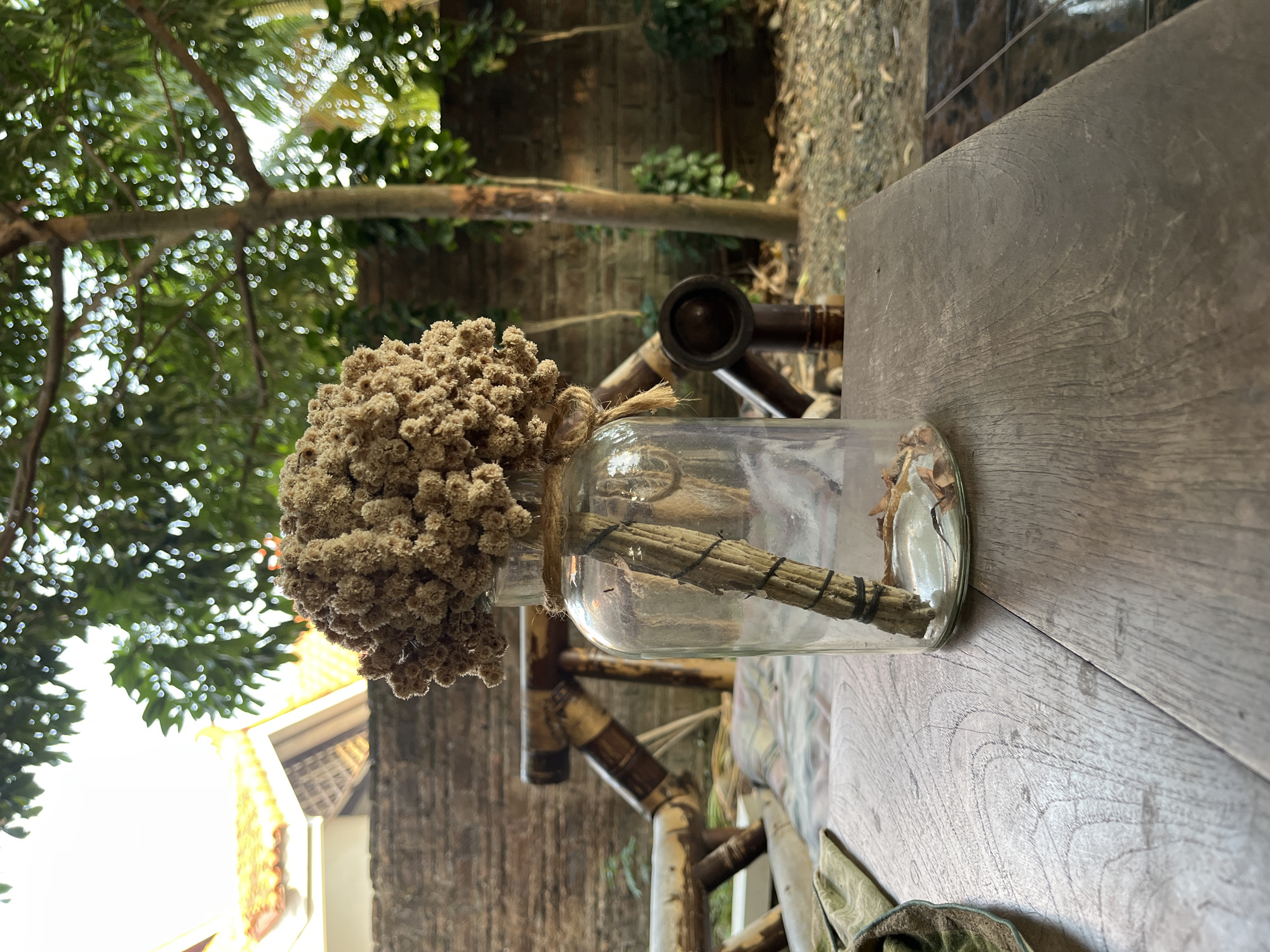 A dried flower arrangement in a clear glass bottle sits on a wooden table outdoors. The bottle is tied with twine and contains a few dried leaves at the bottom. Behind the table, a brick wall is visible, with a rustic wooden bench or chair and greenery. In the background, a tree with green foliage and part of a building with an orange tiled roof are present. The scene suggests a casual outdoor setting, possibly a garden or patio area in Karangkebagusan, Indonesia.