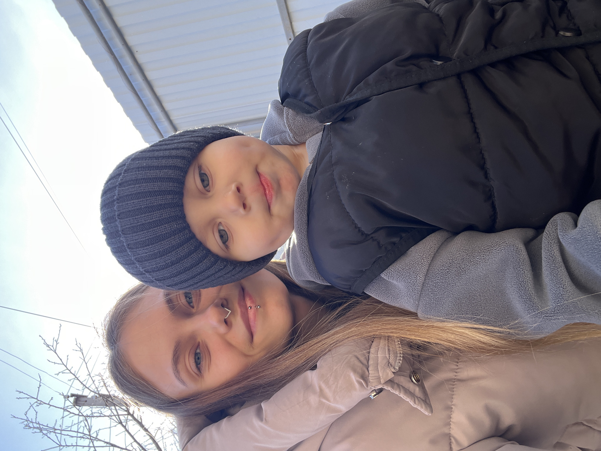 A selfie-style close-up captures two individuals, a child and an adult, from a low angle, likely taken outdoors in Фоки, Russia. The woman has long blonde hair and a nose ring. The child wears a dark-colored knit hat and is dressed in outerwear. Over the woman and the child's head can be seen a silver corrugated awning. In the background, skeletal tree branches and power lines are visible against a bright sky.