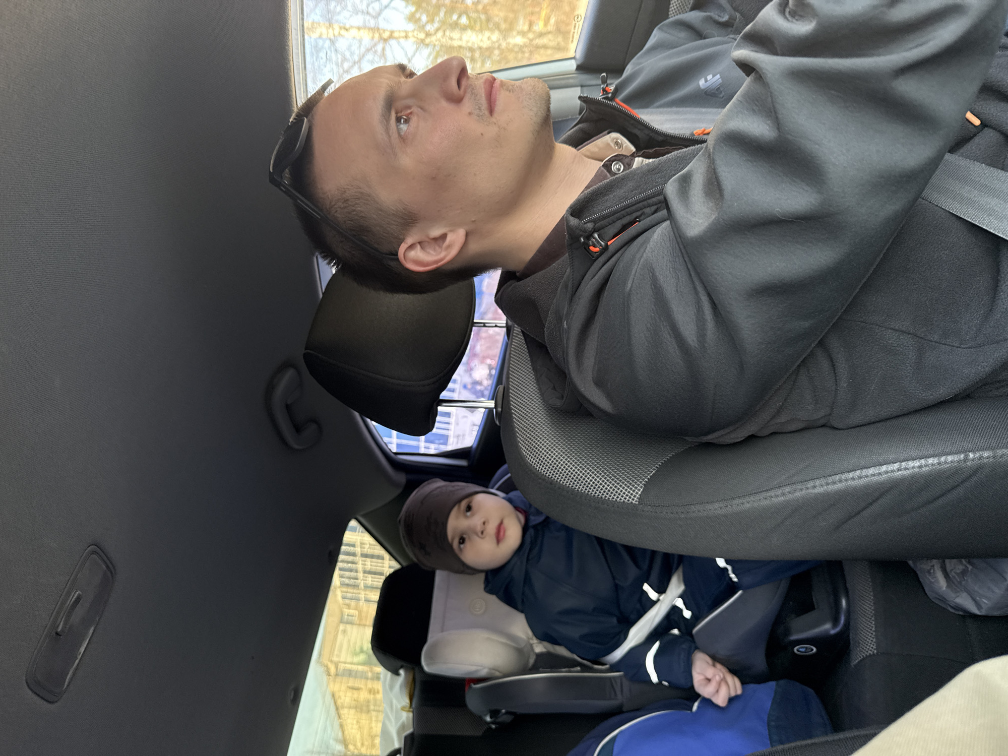 A man and a child are in the back of a car, possibly in transit. The man is in the passenger seat, reclined with his head resting on the headrest and wearing a dark jacket. Sunglasses are on his head. The child is in a car seat, bundled in a jacket and hat. The location might be Chaikovsky, Russia, as indicated by the image request. The background visible through the car window is blurry, with the suggestion of buildings and trees, indicating an urban or suburban environment.
