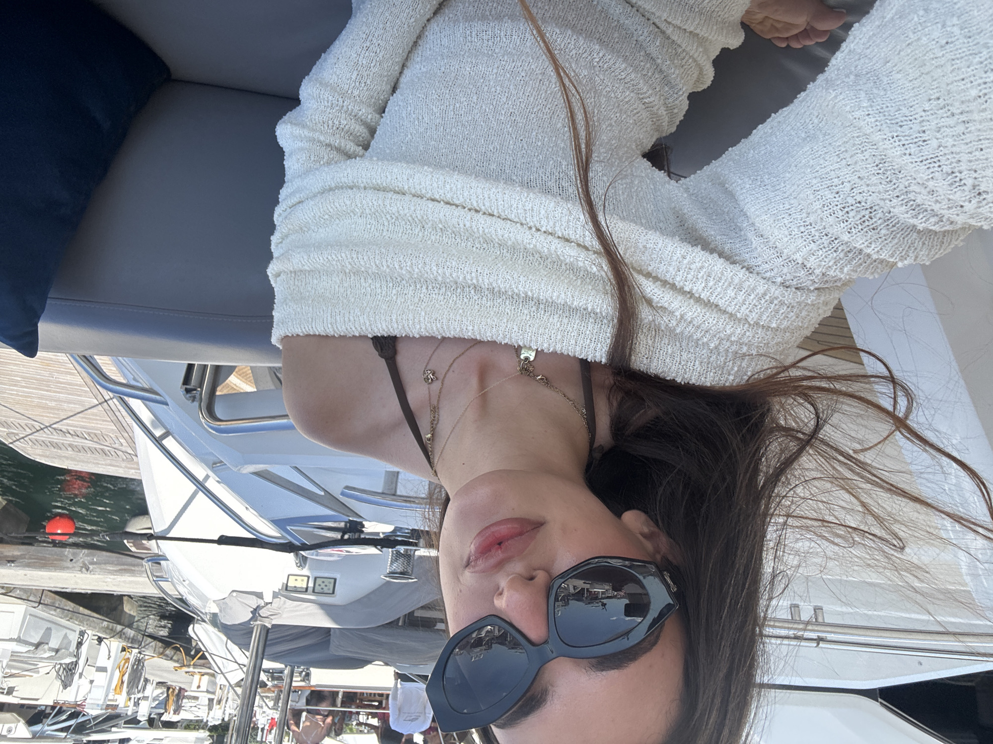 A rotated, high-angle, close-up shot shows a woman lying on a seat on a boat, seemingly in Miami Beach, Florida. She is wearing a white, textured knit outfit and sunglasses. Two delicate necklaces adorn her neck. Her dark hair is tousled. The background reveals other boats and waterfront elements, hinting at a marina environment.