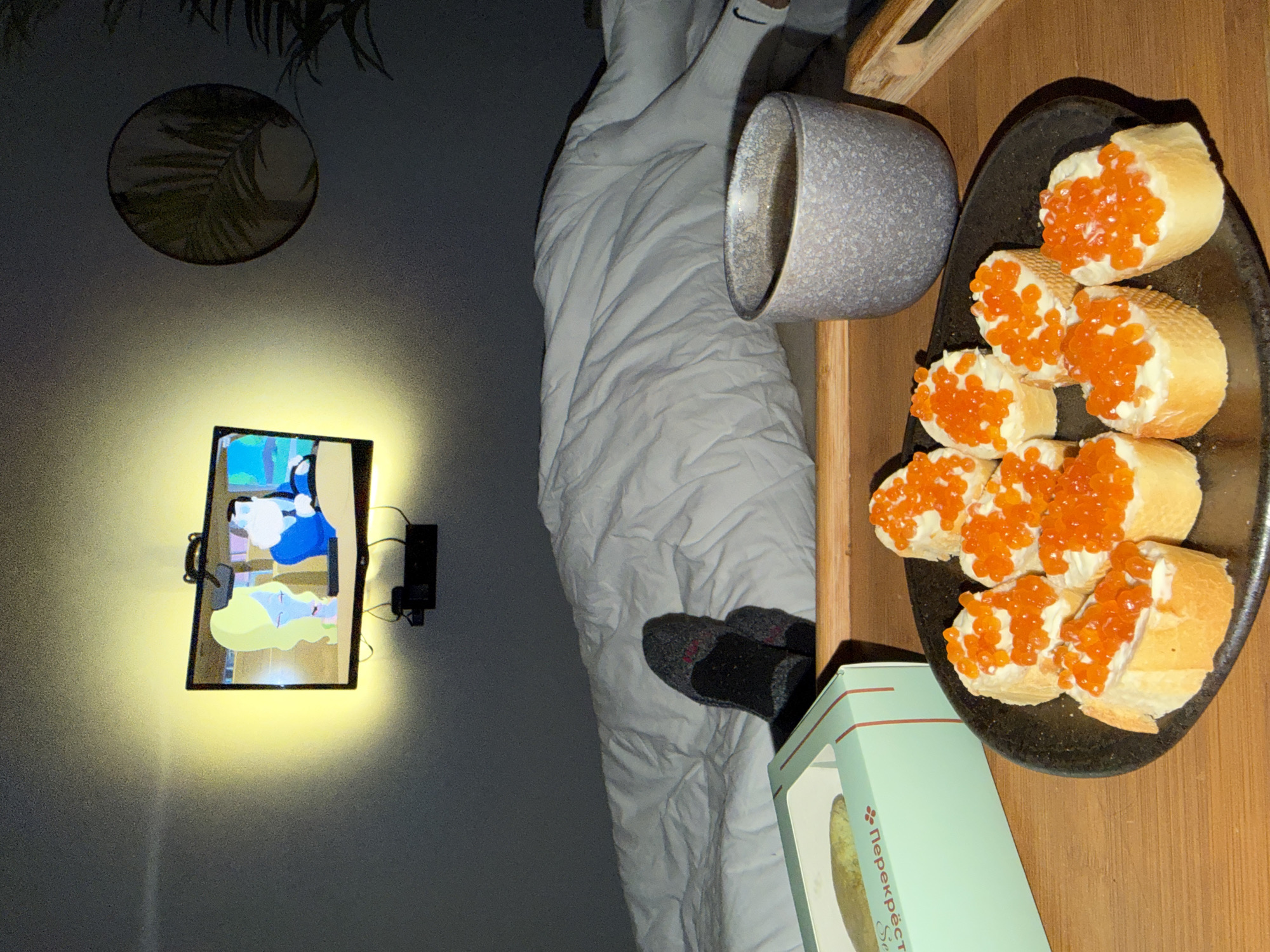The image depicts a cozy, low-light indoor setting, likely a bedroom or living space, during the evening or night. The scene is presented from a slightly elevated, angled perspective.
In the foreground, a person's feet, clad in dark grey socks with red accents, are visible, peeking out from beneath a white, rumpled duvet or blanket. To the right of the person, a wooden tray holds a snack and a drink. On the tray, a black, oval-shaped plate contains several pieces of sliced baguette, each topped with a creamy white spread and abundant bright orange salmon roe (caviar). Beside the plate, a speckled grey mug, possibly holding a beverage, and a light green rectangular box are present. The box prominently features Cyrillic text, reading "Перекрёсток" (Perekrestok), a well-known Russian supermarket chain, and "ИКРА горбуши" (Ikra Gorbushi), signifying Pink Salmon Caviar.
On the wall in the background, a flat-screen television is mounted, casting a warm, bright yellow glow around it. The screen displays an animated image featuring blue and yellow figures. Wires and a black power adapter are visible attached to the wall near the TV. Above the television, a circular wall decoration, possibly a wicker or framed print, shows a design of a palm-like leaf.
The overall activity depicted is one of relaxation, with a person enjoying a snack and watching television. The presence of "Перекрёсток" and "ИКРА горбуши" text confirms the location context of Russia, specifically Khimki. The dim lighting from the room, contrasted with the glow of the TV, emphasizes the evening or nighttime setting and suggests comfort and leisure.