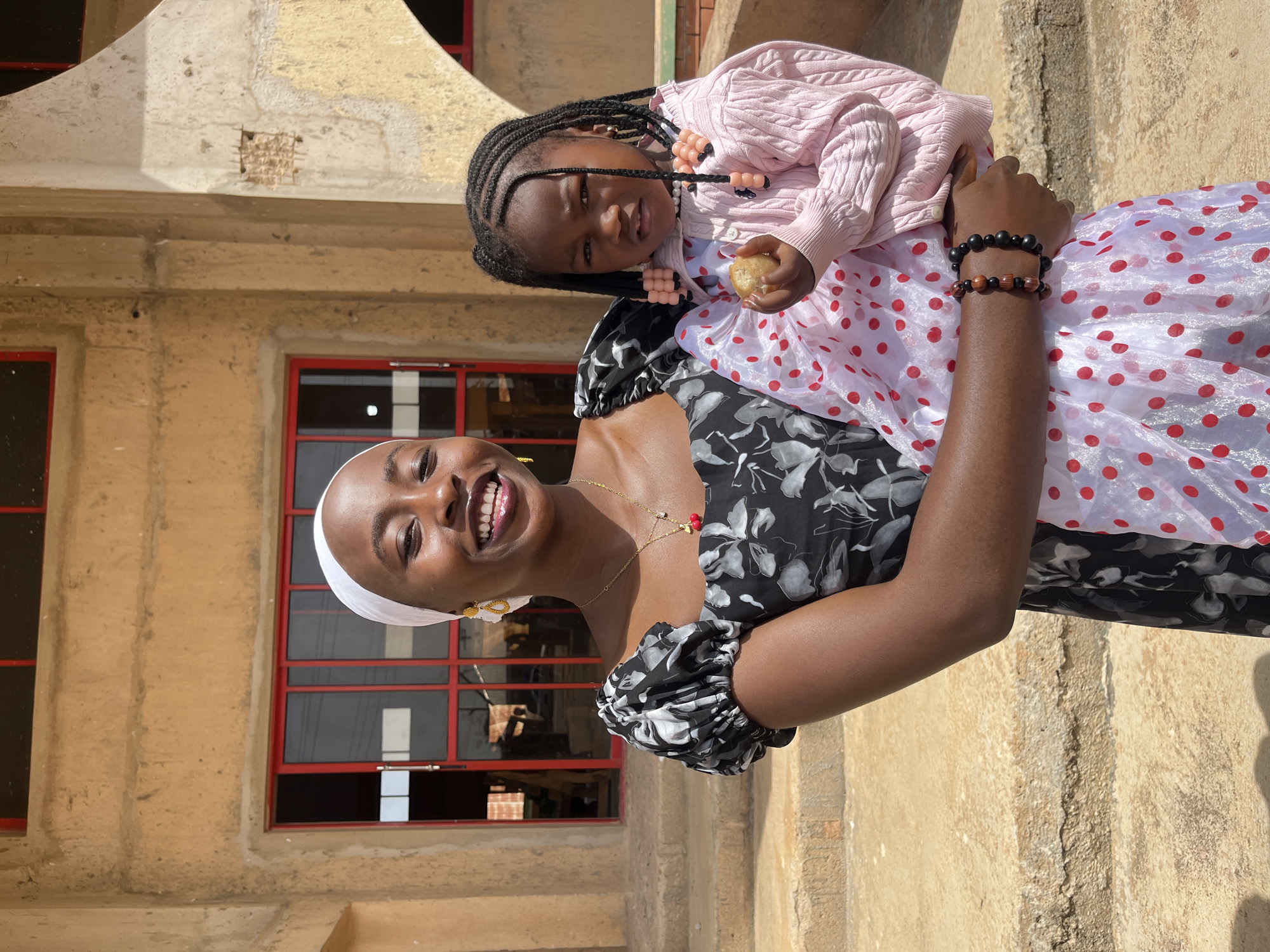 In the city of Bukuru, Nigeria, a young Black woman wearing a white headscarf and a black floral dress smiles at the camera. She is holding a young Black child in her arms. The child is wearing a pink sweater and a white dress with red polka dots, and is holding a piece of food. They are standing in front of a building with beige walls and red-trimmed windows. The ground is concrete and there are stairs leading up to the building.