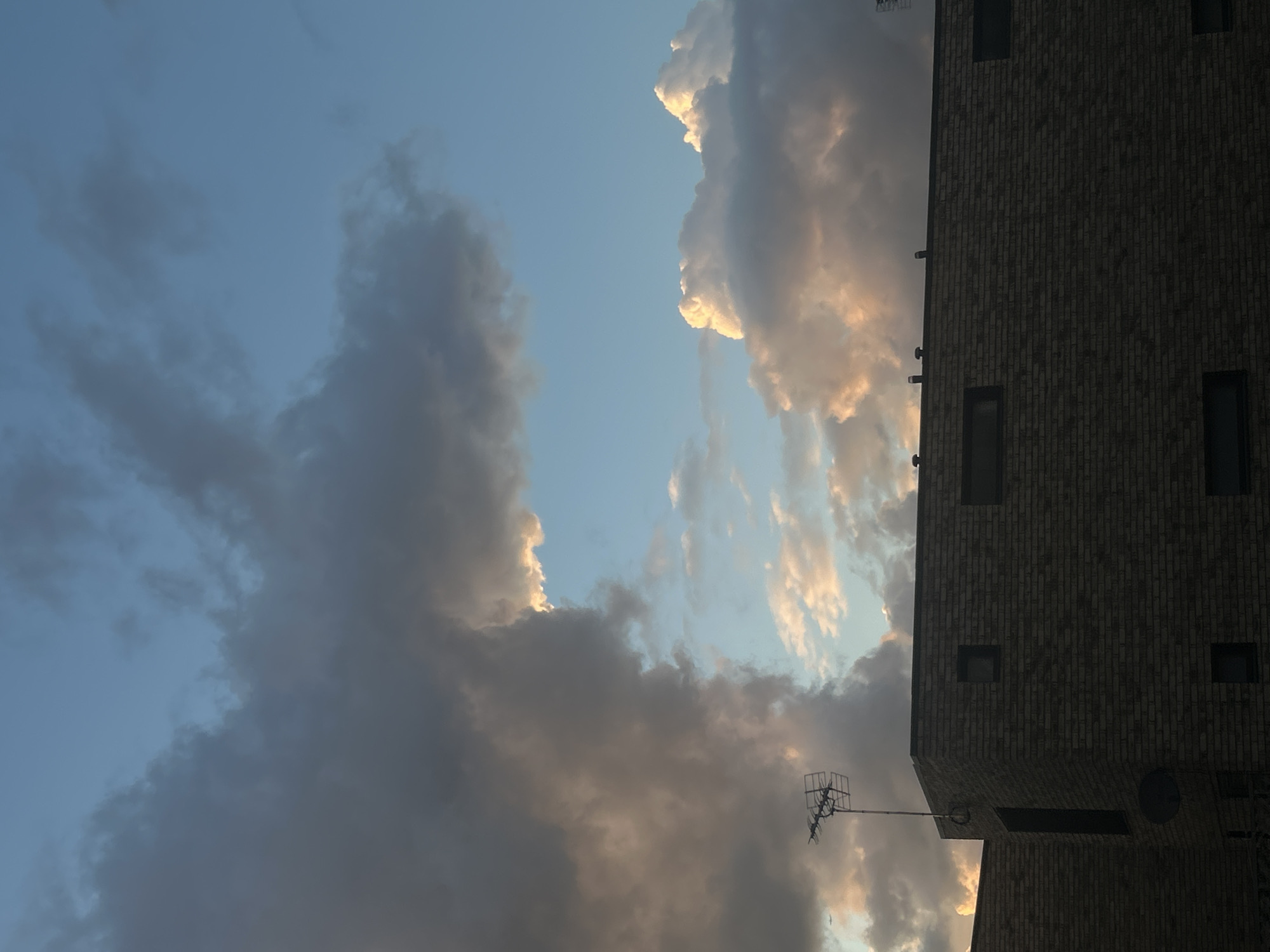 The image shows a cloudy sky next to a brick building in what appears to be the city of London, United Kingdom. The sky is blue with dark and light clouds, some touched with golden light. On the right of the frame, a portion of a brick building with rectangular windows and an antenna is visible.