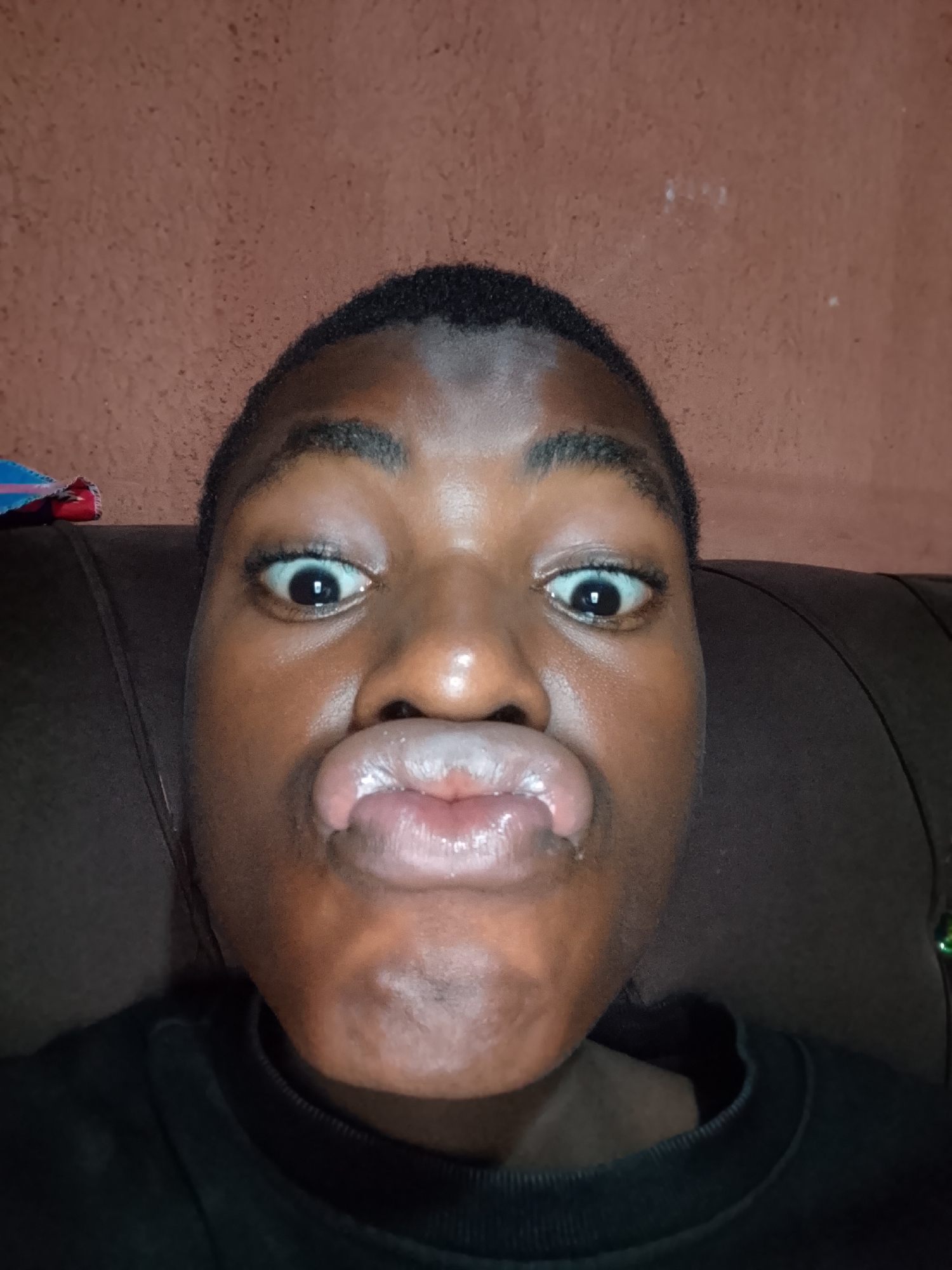 A close-up, front-facing image features an individual with dark skin, short dark hair, and dark eyebrows. The person is making an exaggerated, humorous facial expression, characterized by wide, open eyes revealing much of the whites, and lips puckered into a "fish face" or "kissy face." They are wearing a dark-colored garment, possibly a t-shirt or sweatshirt.
The background shows a dark brown headrest, likely from a couch or chair, immediately behind the person. Further back, there is a reddish-brown wall with a slightly textured appearance. A small, colorful object with blue and red hues is partially visible in the upper-left corner of the frame. The lighting suggests an indoor setting, likely under artificial light. The overall mood conveyed by the facial expression is one of playfulness or silliness. No visible text is present in the image.