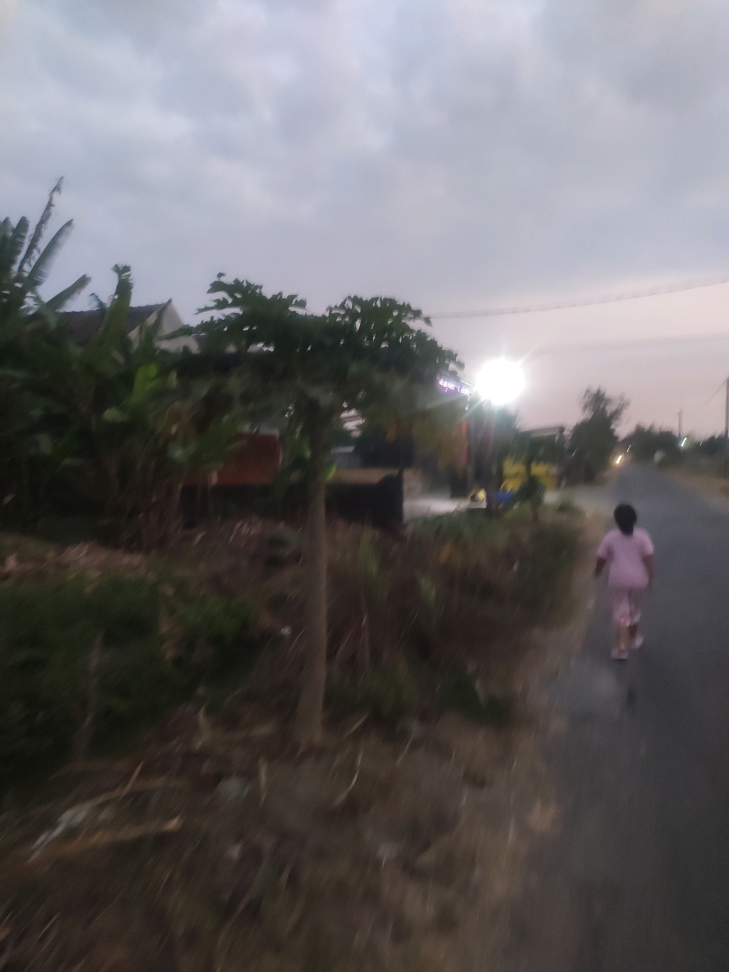 The image depicts a blurry, low-light outdoor scene. In the foreground and mid-ground, there is vegetation including trees and what appear to be banana plants. A road runs diagonally from the lower right to the upper right of the frame. A young girl wearing a pink t-shirt and pink pajama pants is walking away from the camera on the right side of the road. Further down the road, there is a bright light source, possibly a streetlamp, which is overexposed. In the background, there are some buildings with pitched roofs and other foliage. The sky is overcast and dusky, suggesting it is either early morning or late evening. The overall impression is that of a rural or suburban area in Bongkot, Indonesia.
