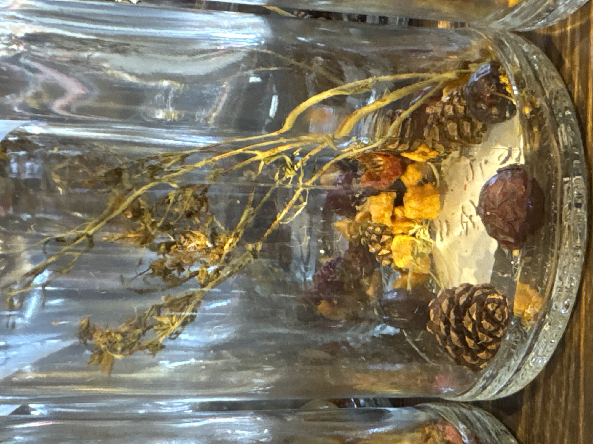 A close-up shot of two glass jars filled with dried botanical elements. The visible contents include dried sprigs, yellow and red flower pieces, and pinecones. The jars appear to be cylindrical, with thick glass and ridges around the top. The background is blurred, but a wooden surface is visible. The image is taken in a natural light setting. The photograph does not depict the city of Kurbaki, Russia.