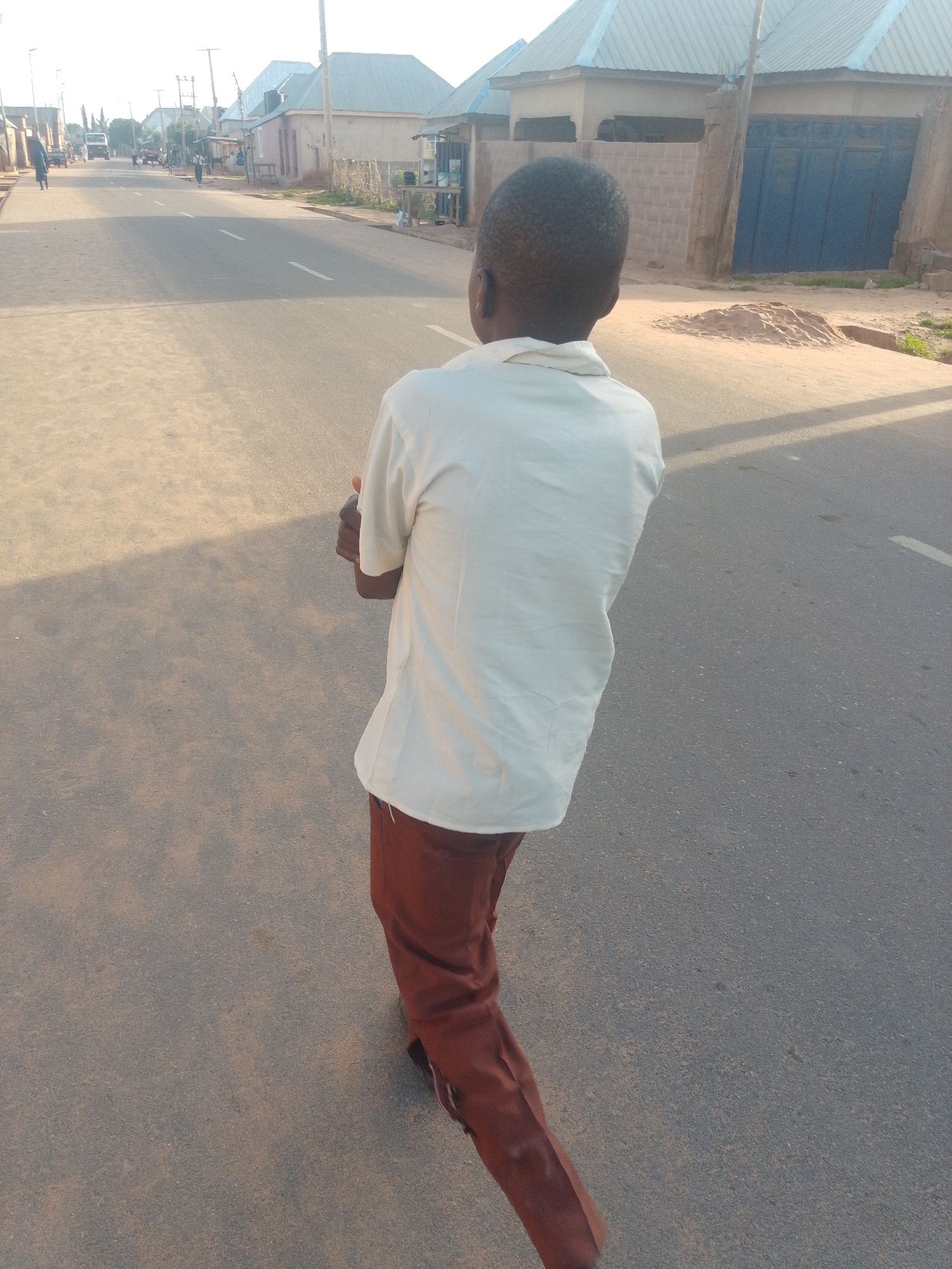 A young boy, viewed from behind, walks away from the camera down a paved street in a residential area. He is wearing a light-colored short-sleeved shirt and brown trousers. His right arm is bent at the elbow, with his hand resting on his chest. The street is lined with buildings on either side, some with corrugated metal roofs. In the background, other people and vehicles can be seen on the street. The lighting suggests it is either early morning or late afternoon.