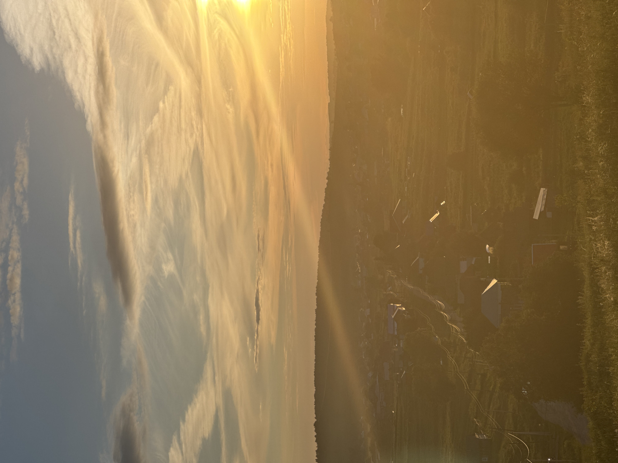 This is an aerial, sideways photograph of a rural landscape at sunset in Baiki, Russia. The image is split into two distinct halves. The left side is dominated by the sky, filled with wispy, golden-yellow clouds that are illuminated by the setting sun, which is positioned high and off-frame to the top left. The sunlight creates streaks and bands of light that stretch across the sky. The right side of the photograph shows a village scene. Several buildings with angled roofs are visible, nestled among trees and vegetation. Winding paths or roads connect the structures. The light of the setting sun casts a warm, golden hue over the entire scene, creating a hazy atmosphere and soft shadows. The overall impression is one of tranquility and the peaceful end of a day. There is no visible text in the image.