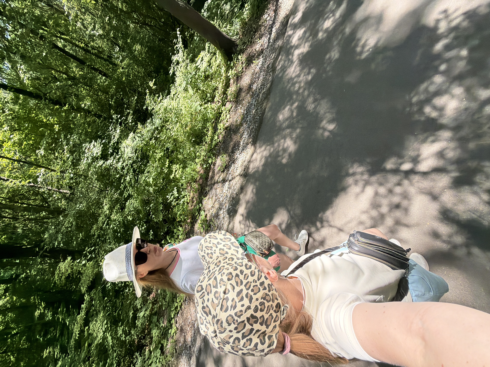 Two individuals, appearing to be women, walk along a paved path in a densely wooded area. The person in the foreground is wearing a light-colored t-shirt, light blue shorts, white sneakers, and a leopard-print baseball cap. They are carrying a black crossbody bag. The second person, further back and to the left, is wearing a white t-shirt, green shorts, white sneakers, and a white fedora hat with a blue and white band. They are also wearing sunglasses. The path is shaded by the surrounding trees, with sunlight filtering through the leaves, casting dappled shadows on the ground. The vegetation is lush and green, typical of a forest environment. No specific landmarks are visible that would definitively place this scene in Khabarovsk, Russia.