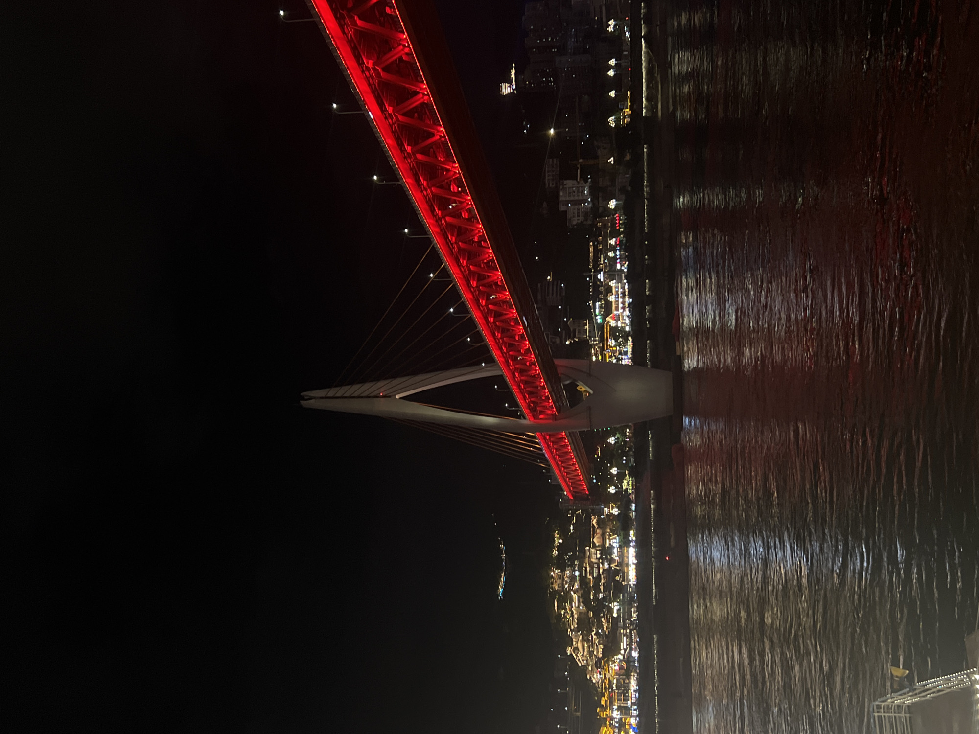 The image depicts a night view of the Hongya Cave bridge in Yuzhong District, China. The bridge is illuminated with bright red lights, highlighting its architectural structure, which consists of a prominent white pylon supporting cables that suspend the red bridge deck. The bridge spans across a dark body of water, likely a river, where reflections of the bridge's lights shimmer on the surface.
In the background, the Hongya Cave complex is visible, with its traditional stilt houses clinging to the mountainside and illuminated by a multitude of warm lights. The overall scene is a vibrant display of urban night illumination, showcasing the blend of modern infrastructure and traditional architecture. The sky is dark, indicating it is nighttime. No people are clearly visible in the image. The vantage point appears to be from another bridge or a boat on the river, looking towards the illuminated city and bridge.