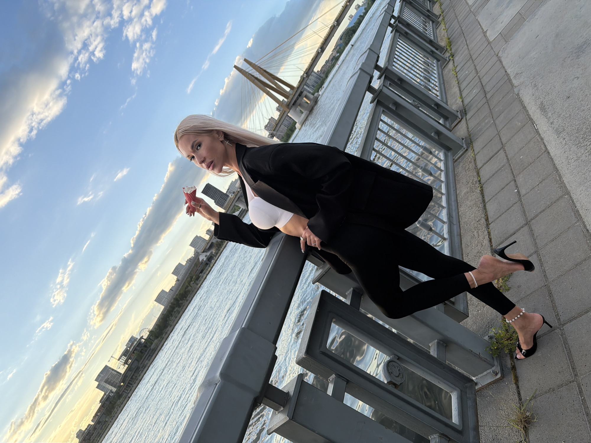A blonde woman with long, straight hair is captured in a stylish pose, leaning on a grey metal railing beside a wide body of water. She is wearing a white ribbed crop top, a black oversized blazer, black leggings, and black high-heeled mules adorned with a pearl anklet on her left foot. In her left hand, she holds a small red ice cream cone. She has a composed expression, looking towards her right, away from the camera.
The scene unfolds outdoors, likely on an embankment or promenade. In the background, a distinctive cable-stayed bridge spans the water, strongly resembling the Millennium Bridge in Kazan, Russia, given the provided location context. A city skyline with various buildings lines the opposite shore. The water surface shows gentle ripples, reflecting the sky and the surrounding structures.
The time of day appears to be late afternoon or early evening, characterized by the warm, golden light of the setting sun illuminating parts of the sky and clouds. The sky is a clear blue with scattered white and grey clouds. The foreground shows a paved walkway made of stone tiles, with a few small green plants growing along the base of the railing.
There is no visible text in the image. The overall impression is one of chic urban leisure during a pleasant sunset.