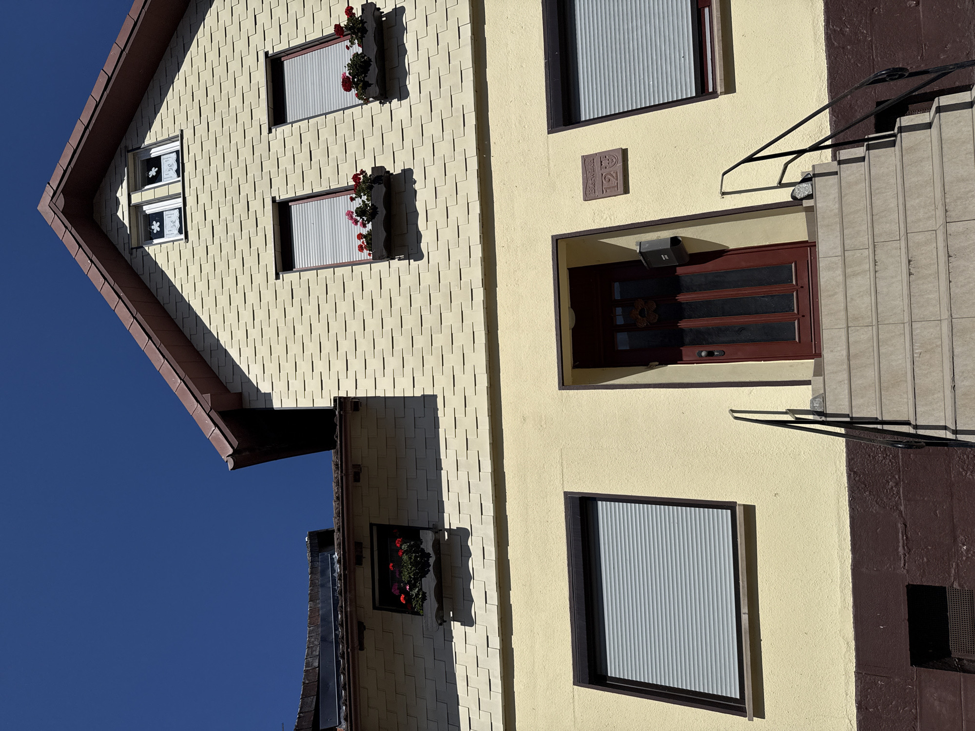 The image shows the facade of a two-story house with yellow siding, a brown roof, and a dark brown base. The house has multiple windows, some with white shutters and others with flower boxes containing red flowers. There is a dark red front door with glass panels, accessible by a set of stone stairs with black railings. A mailbox is mounted next to the door, and a house number plaque reading "12" is visible on the wall above it. The sky is a clear, deep blue. The house is located at Birkenweg 12 in Bad Sachsa, Germany.
