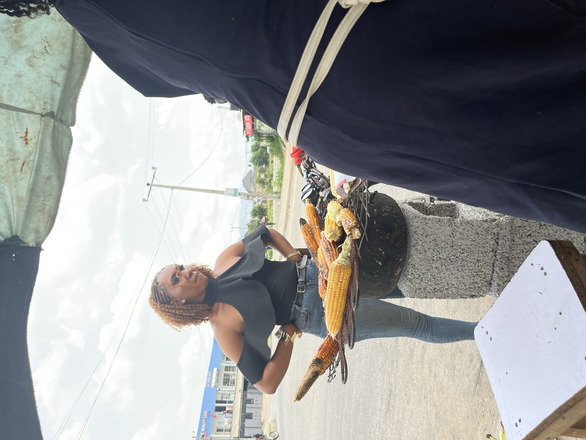 A street vendor stands next to a corn roaster in Abuja, Nigeria. She wears a sleeveless black top, blue jeans, and has golden brown braids. She is posed with a determined expression. The corn roaster is a black, cylindrical device atop a gray stone platform. Several ears of corn, some roasted dark and some lighter yellow, are arranged on the roaster. In the background, a blue building with the words "BEAUTY CARE" is visible. A utility pole with wires stretches across the sky. An awning or partial tent covers the top left portion of the image. Other buildings and greenery are visible in the distance.