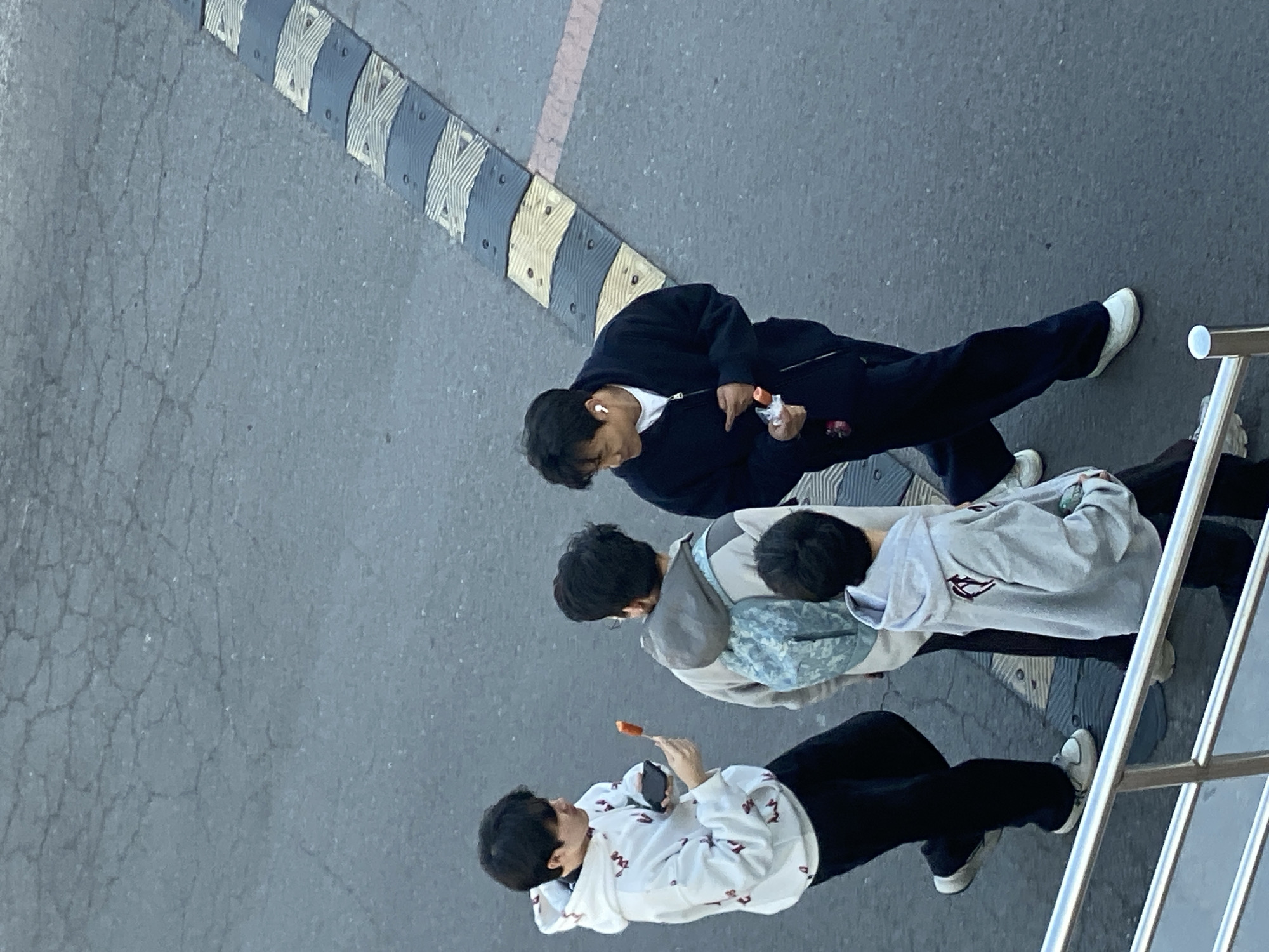 The image shows a street scene in Zhongtun, China. Three individuals are walking along a paved surface. A speed bump is visible on the left. The first person is wearing a dark jacket and white shoes, seemingly holding a snack or wrapper. The second person is wearing a grey hoodie with a light blue camouflage bag. The third person is wearing a white hoodie with red designs and black pants, holding a phone and what appears to be a popsicle. A metal railing is visible on the right side of the image.