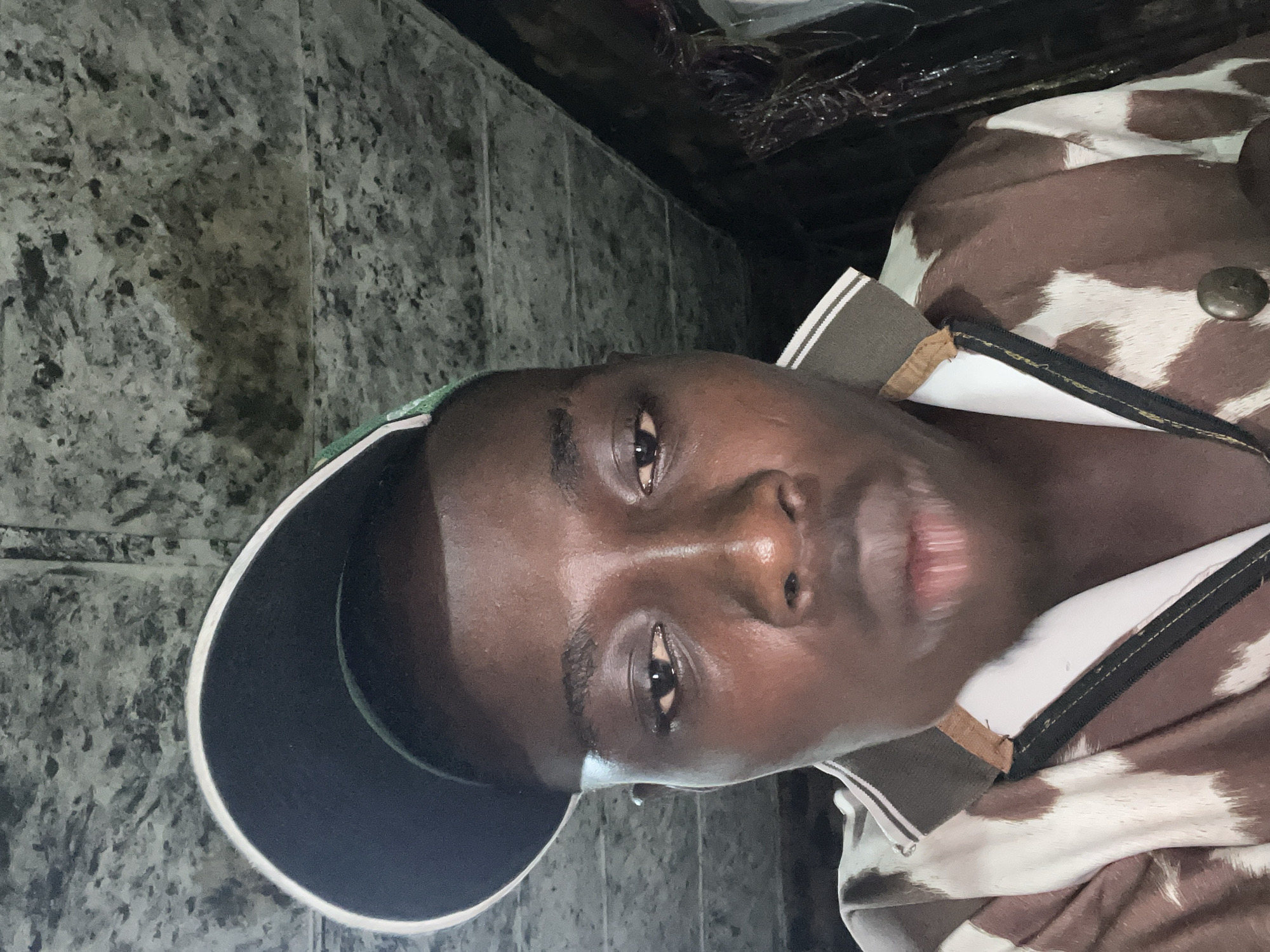 A close-up, low-angle, selfie-style photograph taken indoors. A dark-skinned person wearing a dark baseball cap with a white and green brim and a patterned shirt, possibly with a cowhide design, is the primary subject. The background appears to be a textured, possibly metallic or concrete ceiling, with some areas appearing darker or shadowed. The lighting is dim, creating some glare on the subject's face. The image is cropped closely to the subject, focusing on their face and upper torso. The location in Nkpor, Nigeria is not discernable from the visual information provided in the image.