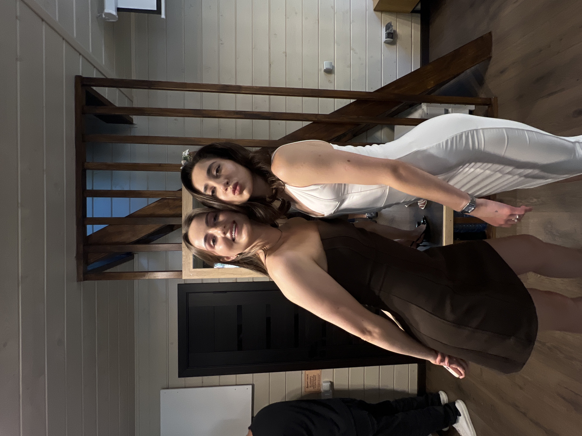 Two women are posing for a photo in a room with wooden walls and a wooden staircase. The woman on the left is wearing a strapless brown dress, and the woman on the right is wearing a white dress. They are both smiling and looking at the camera. The room appears to be indoors, possibly a home or a rented space, given the wooden paneling and the presence of a staircase. The background shows a black door, a mirror, and part of a staircase with wooden railings. There are also some electrical outlets and a light fixture visible on the wall. The floor is made of dark wood. The presence of a whiteboard or projection screen on the left wall suggests it could be a multipurpose room.