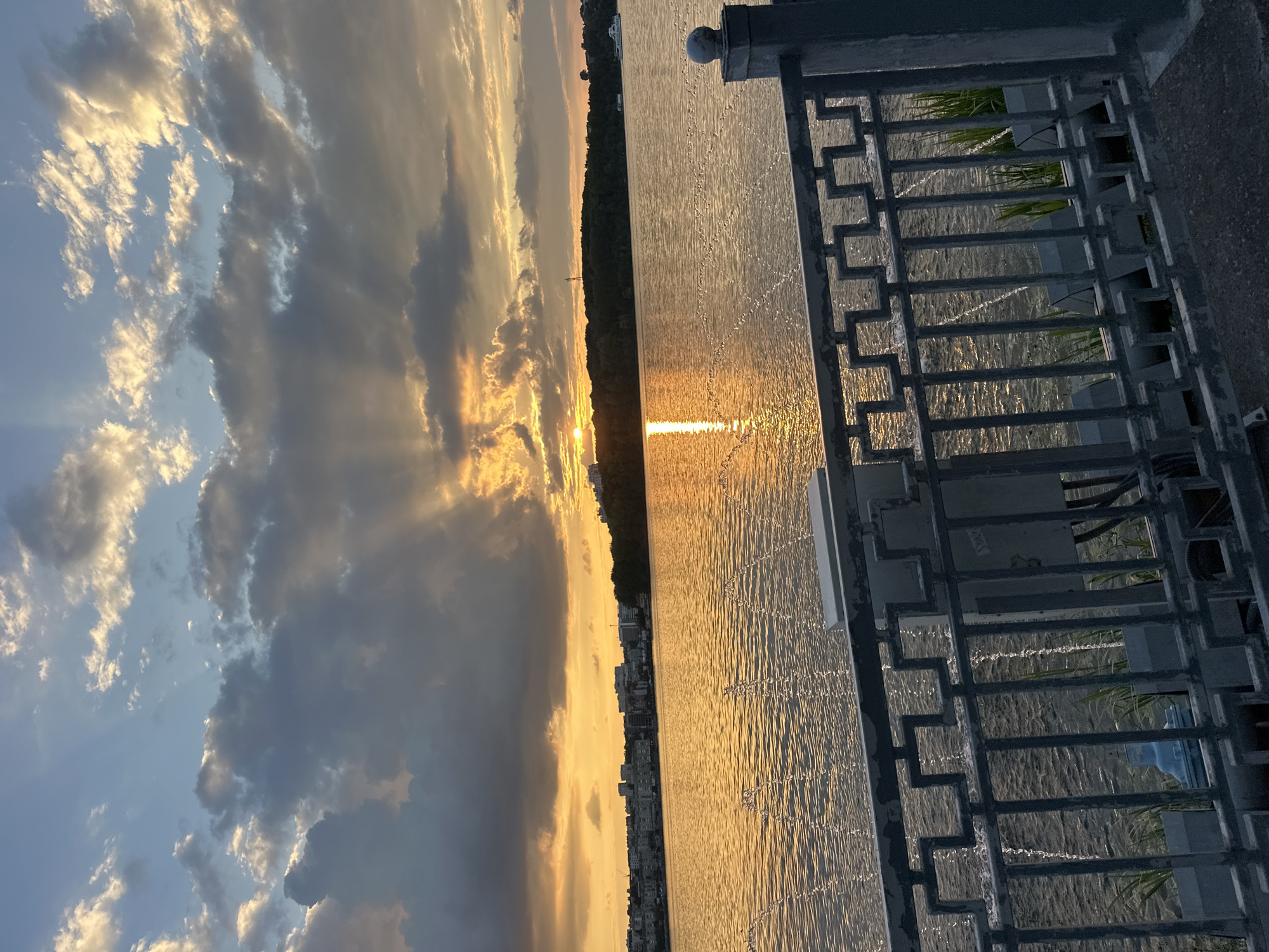 The image captures a sunset over the Ternopil Pond in Ternopil, Ukraine. The sun, a brilliant orb of golden light, is partially obscured by dramatic, dark clouds that are illuminated from behind by its rays, creating a striking visual effect. The sky is a canvas of warm oranges, yellows, and grays, with shafts of light piercing through the clouds.
The surface of the pond reflects the vibrant colors of the sky, creating a shimmering, golden expanse. Small, fountain-like water jets are visible, breaking the surface of the water and adding texture to the scene. In the distance, the city skyline of Ternopil is visible, a collection of buildings nestled against a tree-lined shore. The cityscape appears muted and silhouetted against the bright sky.
In the foreground, a dark metal railing with a geometric design runs horizontally across the image, partially obscuring the view of the pond and the city. Behind the railing, some greenery and electrical boxes are visible. The overall atmosphere is one of natural beauty and urban tranquility, bathed in the warm glow of the setting sun.