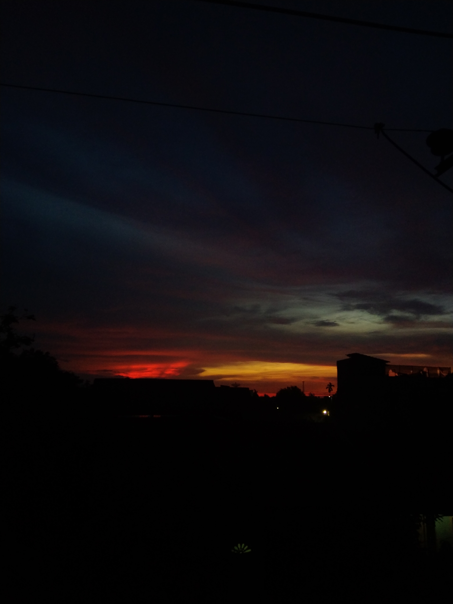 A dusk or dawn landscape in Bireuenmeunasahdayah, Indonesia. The sky transitions from dark blue at the top to vibrant red and orange hues at the horizon. Silhouetted buildings and trees occupy the lower portion of the image. A few utility lines are visible against the sky. A few distant streetlights can be seen. The foreground is dark.