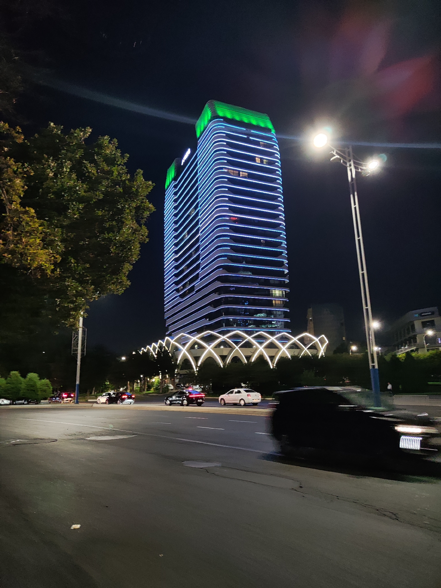 The image depicts a nighttime scene in Tashkent, Uzbekistan, with a prominent modern skyscraper illuminated by blue and green lights. The skyscraper, which appears to be a hotel or office building, has a distinctive wavy facade and is topped with green accent lighting. In the foreground, a street with several cars is visible. One black car in the right foreground is blurred due to motion. On the left side of the street, there's a tree and a streetlamp. In the mid-ground, a decorative archway structure with white lights spans across the scene, connecting to the base of the skyscraper. The overall environment suggests an urban setting with well-lit infrastructure.