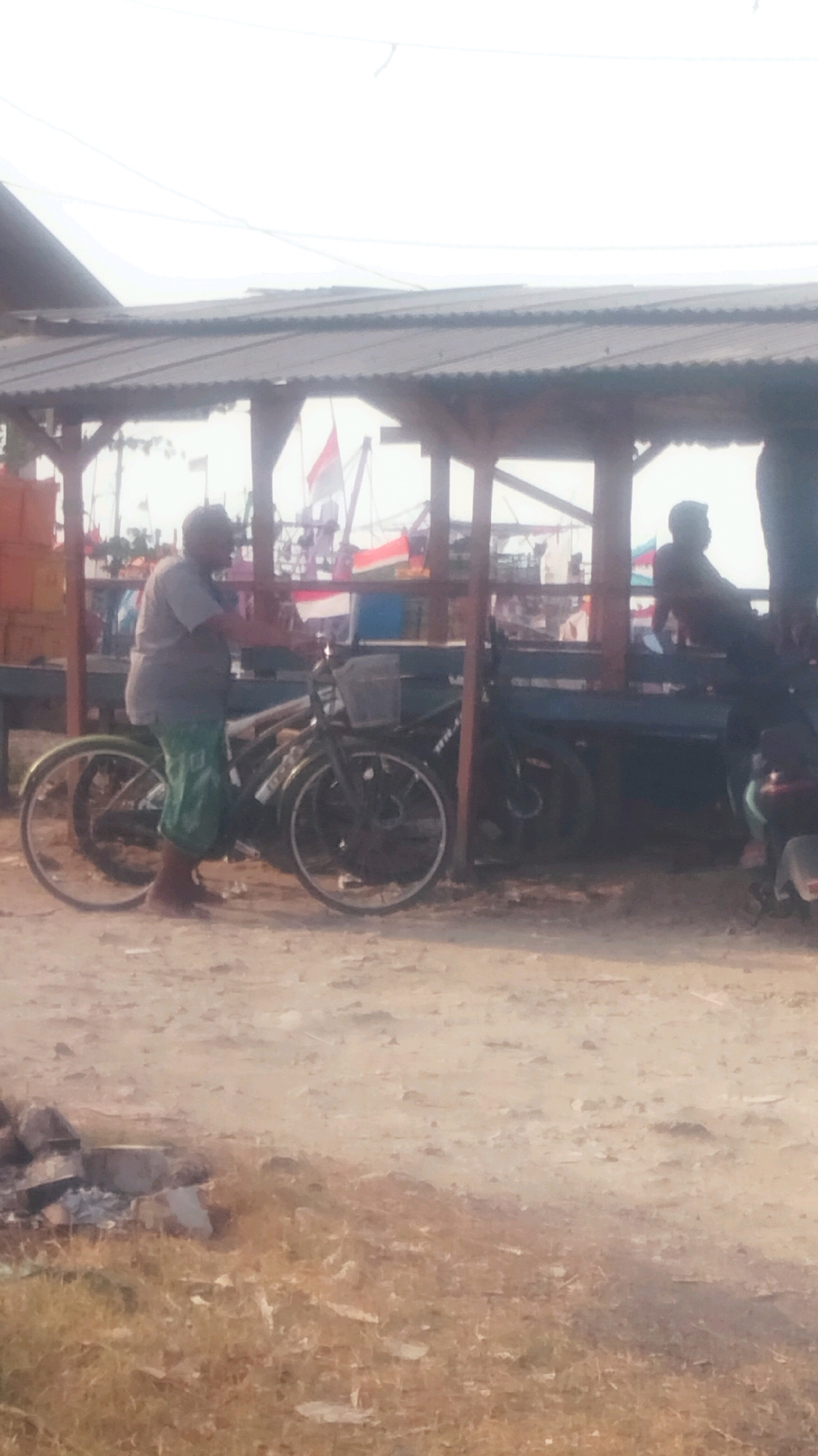 A man stands next to two bicycles under a tin-roofed structure in Palang, Indonesia. The man, wearing a grey t-shirt and patterned green shorts, is barefoot and appears to be interacting with the bicycles. The bicycles are dark-colored, one with a front basket. Behind the structure, several flags are visible, including red and white ones, suggesting a possible cultural or national display. The ground is a dirt and gravel surface with some dry grass and scattered debris. The overall atmosphere suggests an outdoor market or roadside stall.