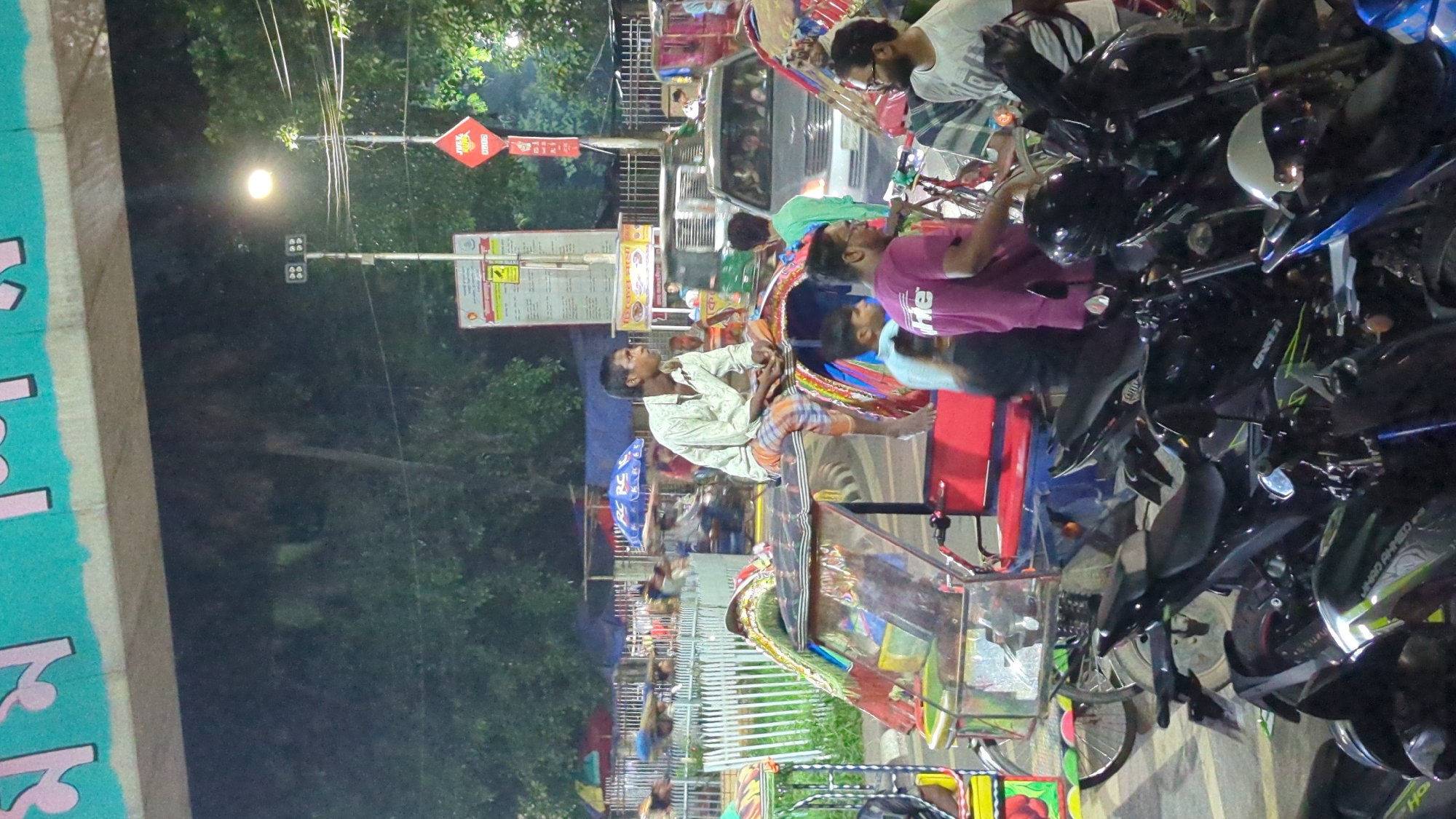 In the city of Dhaka, Bangladesh, a busy street scene unfolds at night. A man is seated on the roof of a colorful auto-rickshaw, looking to his left. He is wearing a light-colored collared shirt and plaid pants. In the foreground, several parked motorcycles are visible. Behind the auto-rickshaw, other vehicles, including a car and more auto-rickshaws, are on the road. People are also seen in the background, some with umbrellas. Overhead, a bridge or elevated structure is visible, with a large green sign displaying Bengali text. Streetlights illuminate the area, contributing to the nighttime ambiance.