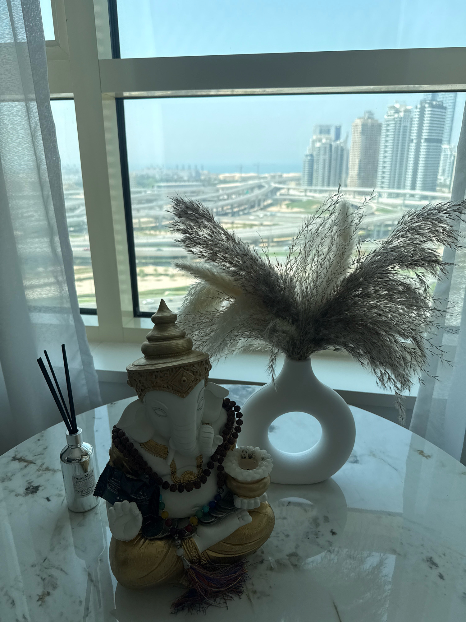 This is a medium shot, eye-level, indoor photograph of a table with decorative items, set against a backdrop of a cityscape viewed through a large window. The table has a white marble top with dark grey veining. On the table, to the left, is a small, clear glass bottle with a silver cap, containing several black diffuser reeds. Next to the bottle is a statue of the Hindu deity Ganesha, depicted in a seated position with a white body, gold accents, and ornate beaded necklaces. To the right of the Ganesha statue is a modern, white ceramic vase with a circular opening in the center, filled with dried ornamental grasses. The window behind the table offers a panoramic view of Dubai, United Arab Emirates, featuring a dense collection of modern skyscrapers, elevated highways with multiple lanes, and a hazy horizon where the city meets the sea. White curtains are visible on the left and right sides of the window, partially obscuring the view. The lighting is natural, coming from the window, and casts soft shadows on the table.