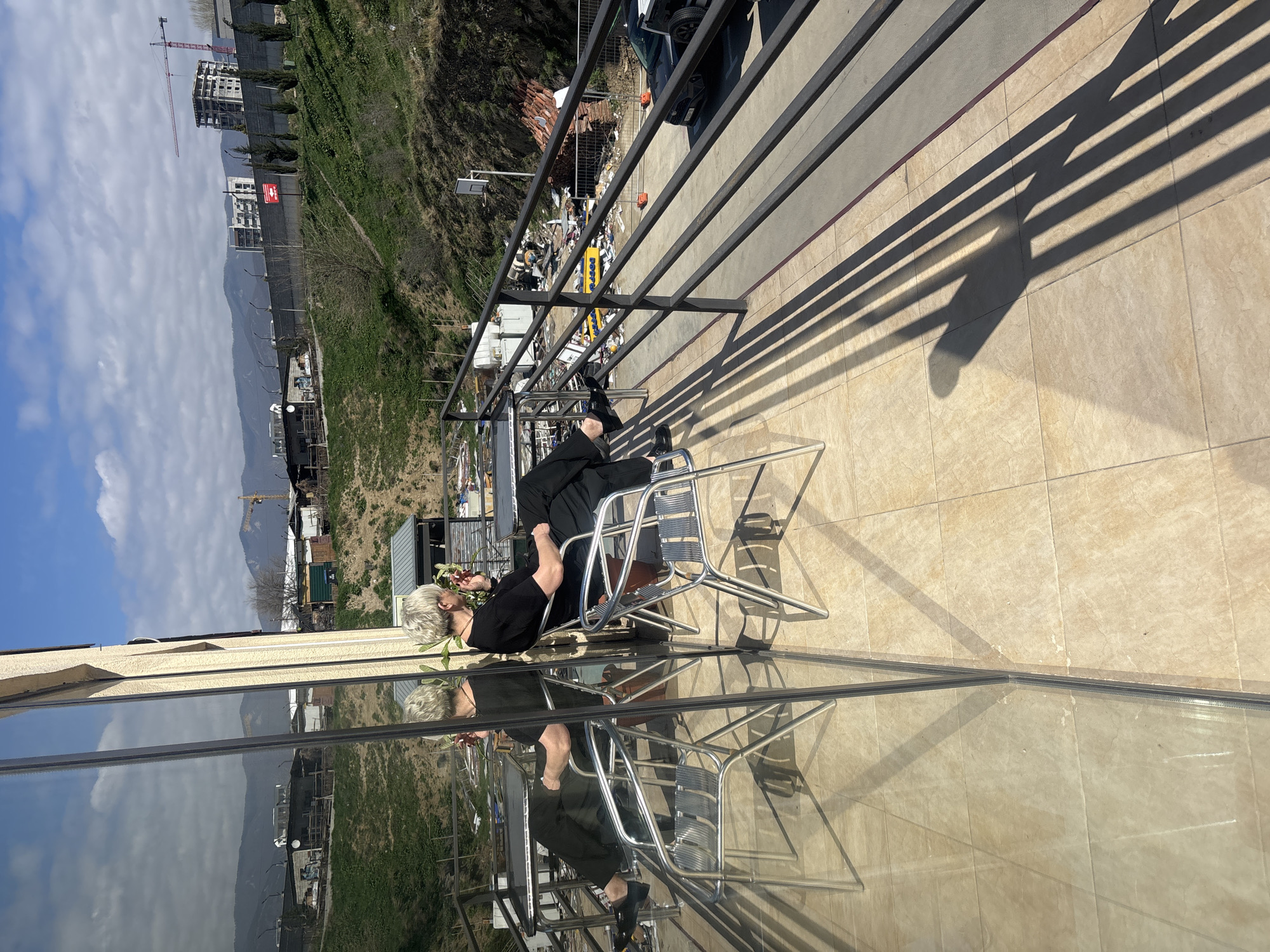 A person with short, gray-blonde hair sits on a silver metal chair on a tiled balcony in Tbilisi, Georgia, reflected in a glass wall. The person is wearing a black shirt and pants. A small flowering plant is in their left hand. The balcony has a black railing that casts striped shadows onto the tile. The balcony overlooks a cityscape with hills, buildings, and construction cranes. Debris and discarded items lie below the balcony. A car is parked nearby. The sky is visible, with patches of blue and some clouds.