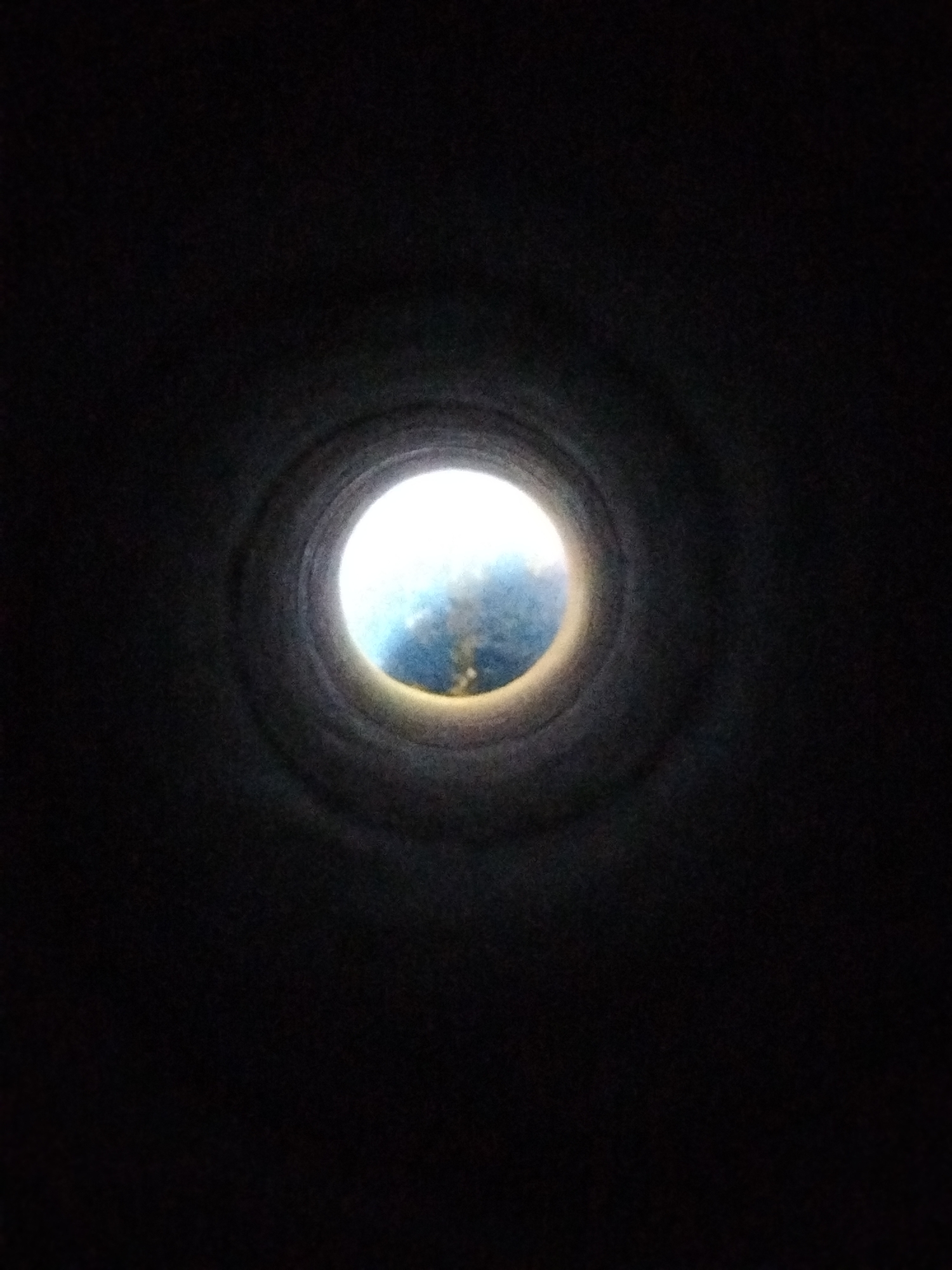 The image shows a circular opening looking out into a blue and white sky, possibly with clouds. The opening appears to be the end of a dark, cylindrical tunnel or pipe. The tunnel walls are textured and cast a shadow, creating a spotlight effect on the view beyond. The overall impression is one of looking out from a confined space into the open. Without additional information or context, it is not possible to determine the specific location in Kremenchuk, Ukraine.
