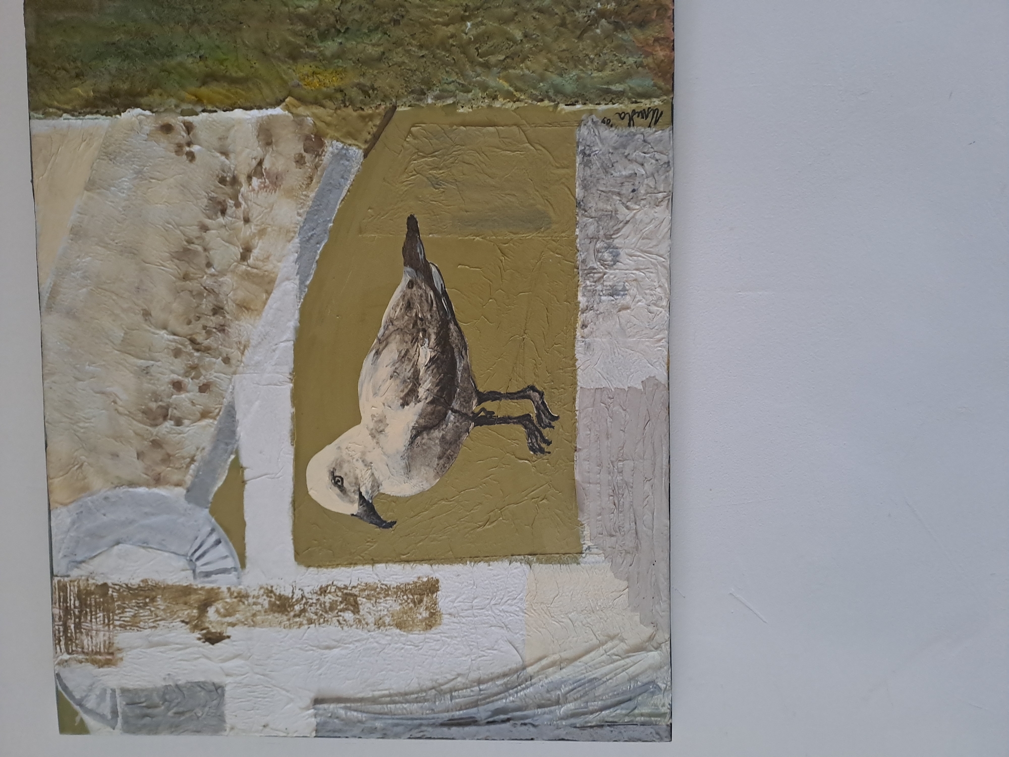 The image shows a square artwork hanging on a white wall. The artwork itself is a mixed media collage or painting with textural elements. The upper left corner features a patch of green and brown textured material suggesting landscape. A central, olive-green square contains an image of a bird in a dive or swooping pose. The bird is rendered in muted tones of white, gray, and brown. The surrounding areas of the artwork consist of layered textures in creams, whites, and browns with hints of metallic silver. A signature is visible in the top right corner of the piece. The artwork appears to be hanging indoors, on a plain white wall. There is no information in the image to suggest it is located in Veliko Tarnovo, Bulgaria.
