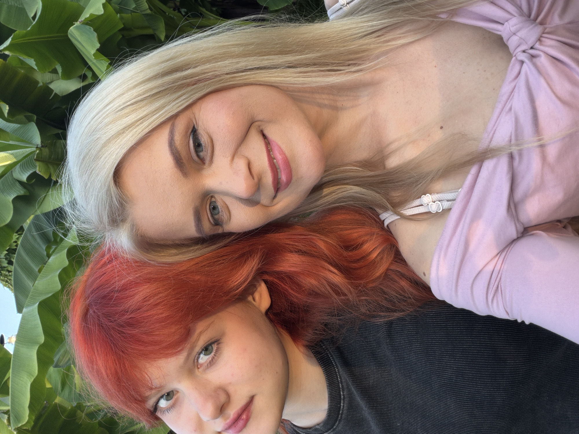 The media file contains a close-up, overhead shot of two young women posed together. The woman in the foreground has vibrant red hair and is looking upwards, with her eyes open. She is wearing a dark, textured t-shirt. The woman behind her has long, blonde hair and is also looking towards the camera, with a slight smile. She is wearing a light pink, long-sleeved top with a knotted detail. Both women appear to be in good health and are smiling. The background is dominated by large, lush green leaves, suggesting an outdoor setting with tropical or subtropical vegetation. The lighting indicates it could be daytime, possibly late afternoon given the soft light. There is no visible text in the media file. The scene is intimate and friendly, with the two women leaning into each other. Based on the provided location context, the setting could be a garden or park in Sochi, Russia, known for its warm climate and abundant greenery.