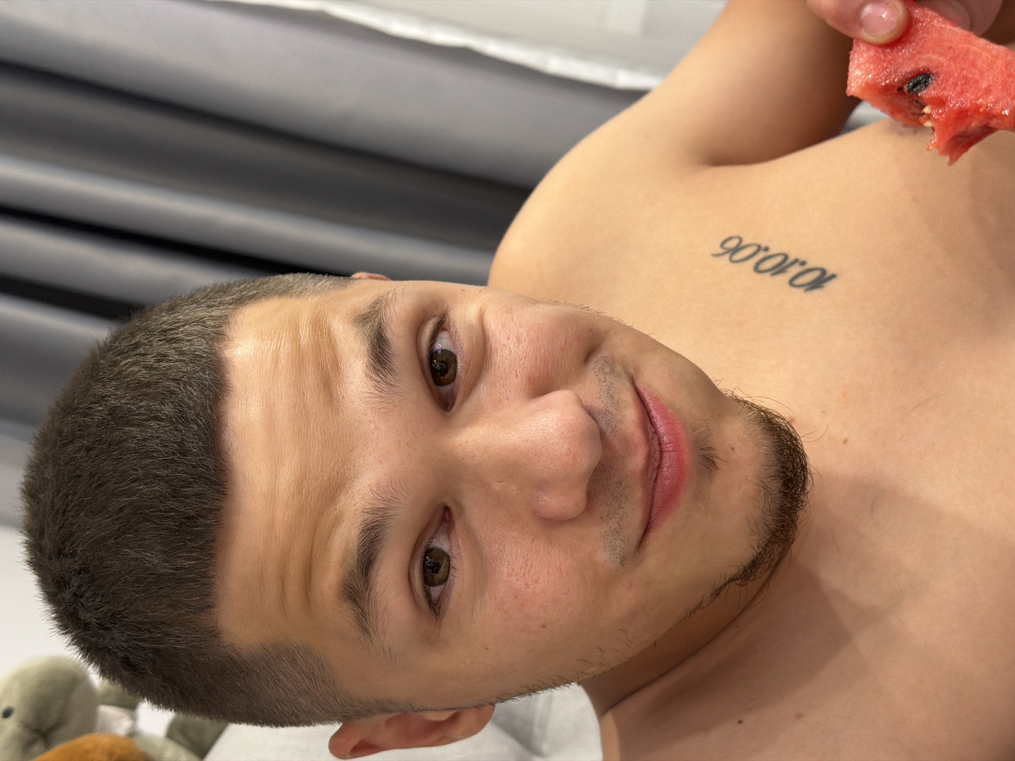 The image shows a close-up of a shirtless man with short dark hair and a goatee. He has a tattoo on his chest that reads "10.10.06". He is holding a slice of watermelon and appears to be eating it. The background consists of gray curtains. The image does not provide any information about a location in Sofia, Bulgaria.