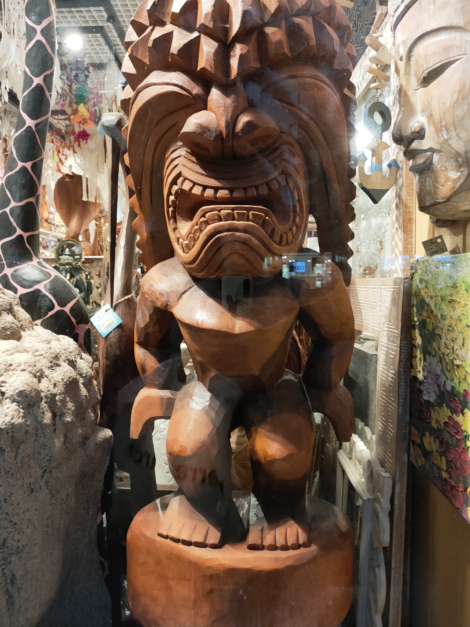 In an indoor market stall in Sochi, Russia, a large wooden tiki statue is prominently displayed. The statue depicts a fierce face with a wide, open mouth filled with carved teeth, and a headdress of sharp, angular protrusions. The body is muscular and crouched. To the right of the tiki statue, a light-colored wooden carving of a serene human face, possibly Buddha, is visible. The background is cluttered with various decorative items, including a black and pink patterned pole on the left, and colorful fabric or artwork on the far right. Reflections in the glass show a ceiling and possibly other market stalls.