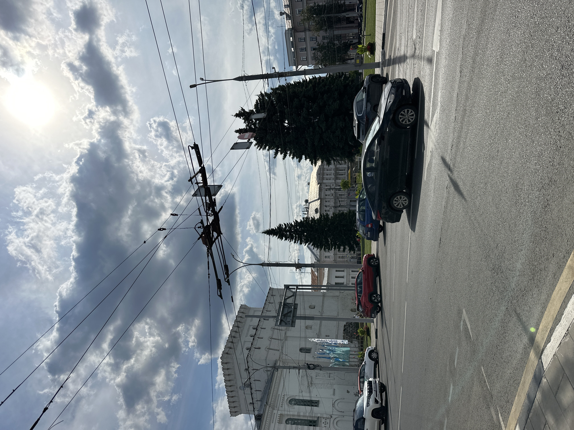 A street scene in a city, likely Putyatino (Semivragovskaya S/A), Russia, on a partly cloudy day. Overhead wires for a trolleybus system crisscross the frame. On the left, a white, fortress-like building with crenellated battlements and arched windows stands prominently. In front of it, several cars are visible on the road: a white SUV, a red SUV, a dark sedan, and a dark blue sedan. A tall evergreen tree is positioned behind the cars, near a lamppost. Further in the background, various multi-story buildings with classical architectural elements line the street. The sky is bright with the sun peeking through scattered clouds, casting shadows on the asphalt road and vehicles. The road has lane markings, including a crosswalk.