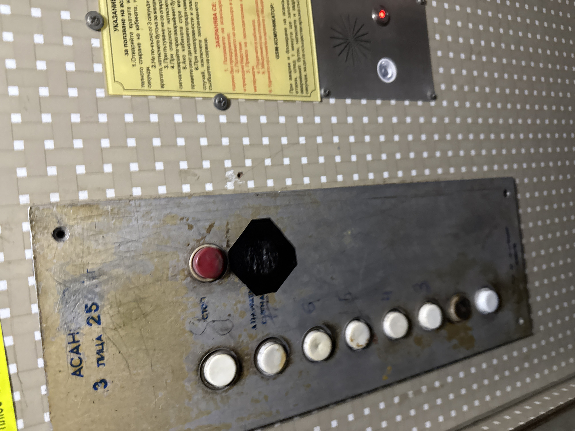 The image shows a close-up view of an old elevator control panel. The panel is metallic, likely made of stainless steel, and shows signs of wear and tear, including scratches and discoloration. The panel features several white push buttons labeled with floor numbers, a red emergency stop button, and a black hexagon-shaped element, possibly a speaker or microphone. Handwritten in blue ink next to the panel are the words "ACAH 3 лица 25," which could be instructions, weight limit (in лица), or identifying marks. Above the panel, two yellowed, laminated signs are affixed, containing safety instructions written in Bulgarian. Above these, there is a small, metal plate with a red light. The control panel's condition suggests it is in an older building with outdated or unmaintained elevator equipment.