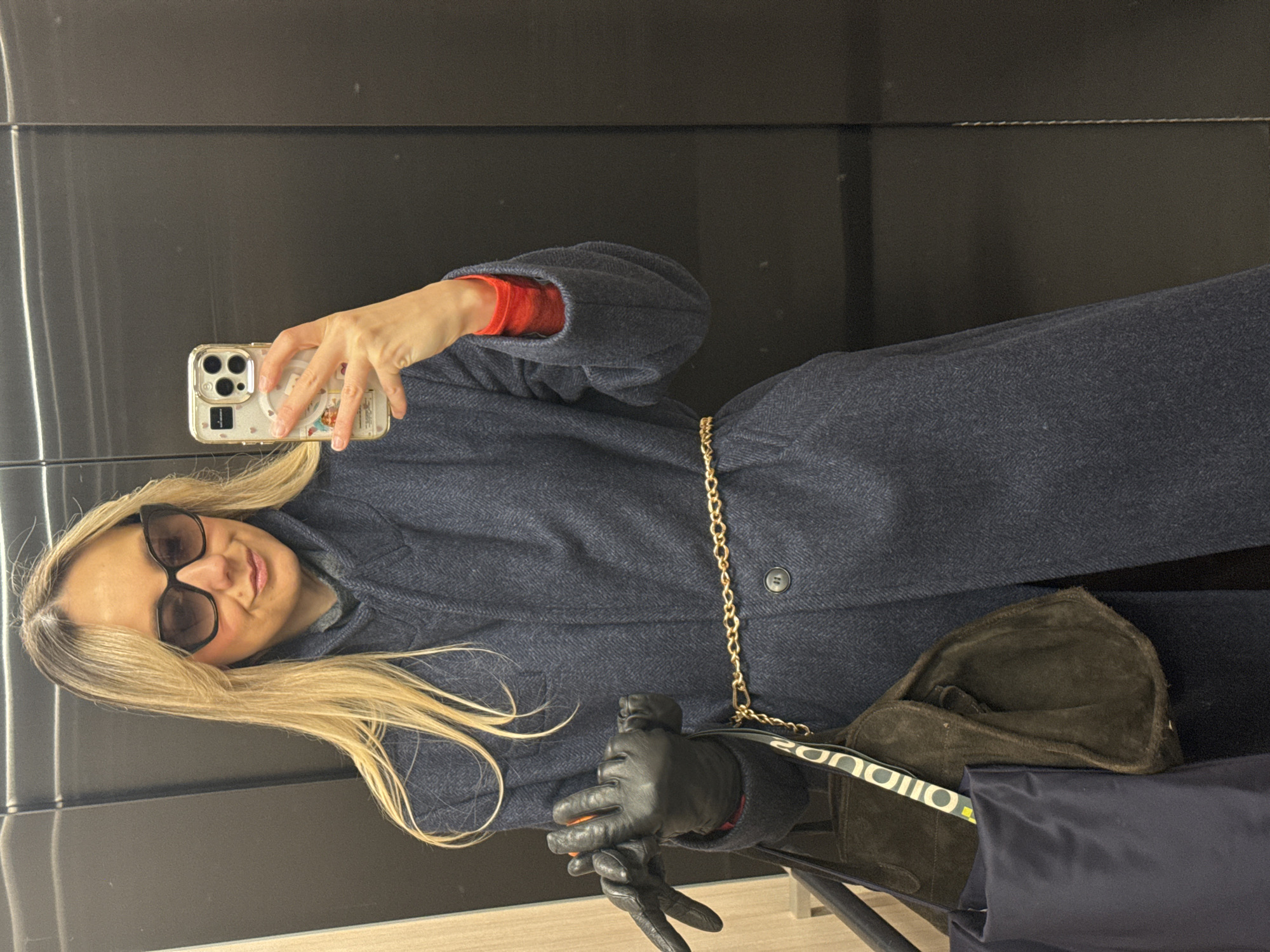 A woman takes a selfie in a reflective surface, likely an elevator or a large mirror, in what is contextually Riga, Latvia. She has long, blonde hair and wears dark sunglasses, with a slight, relaxed smile.
She is dressed in a dark blue, possibly herringbone-patterned, long coat, under which a bright red long-sleeved top is visible at the sleeves and collar. A thin gold chain is draped around her waist, serving as a decorative belt. She wears black leather gloves on both hands.
In her right hand, she holds a smartphone, specifically an iPhone with multiple cameras, protected by a clear case adorned with small, colorful elements. Her left hand, also gloved, rests near a brown suede bag. A portion of the bag's strap is visible, displaying partially discernible text that reads "EON D U O" or similar. Another dark blue fabric bag or pouch is visible next to the suede bag. The background is a uniform dark, glossy, and reflective surface.