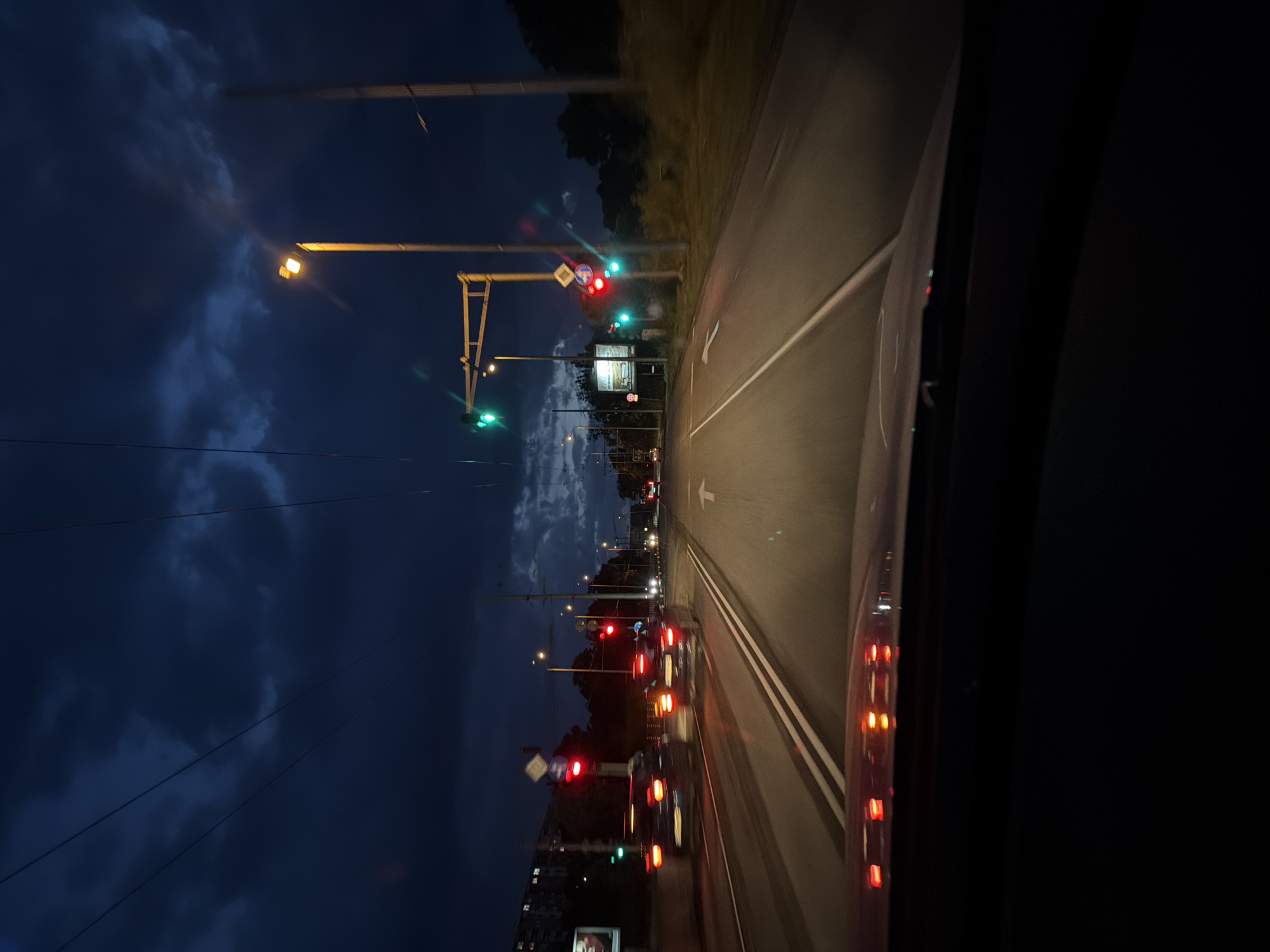 The image depicts a view from inside a vehicle, looking out onto a city street at night. The location is likely Burgas, Bulgaria. The street is illuminated by streetlights and traffic signals, many of which are red. Traffic is visible, consisting of several cars, many with their tail lights illuminated, indicating they are stopped or moving slowly. There is a road with white lines. Some building silhouettes are visible on the left side of the frame, and a darker area, possibly trees or parkland, is visible on the right. The sky is a deep blue, indicating twilight.