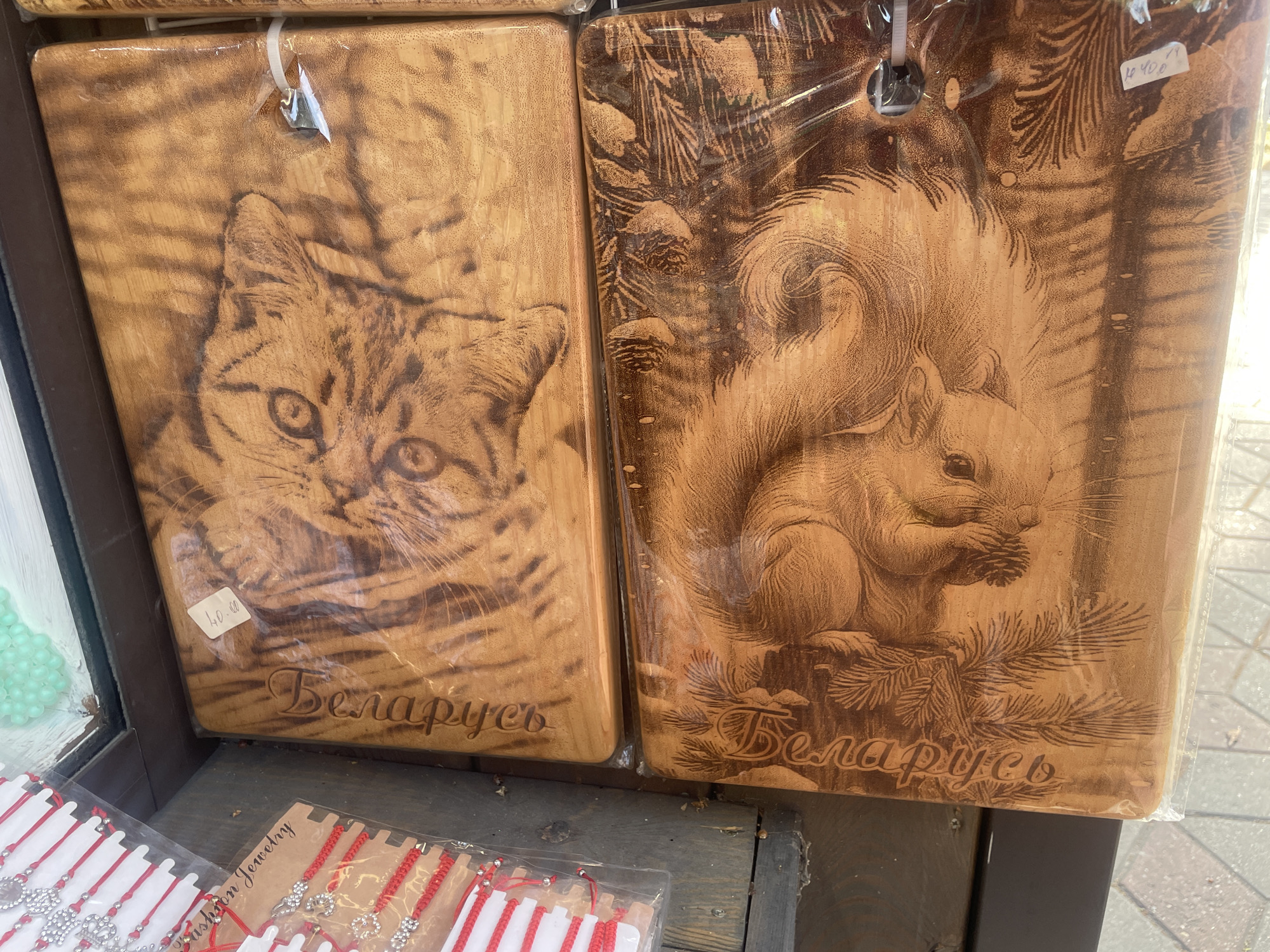 The image displays two wooden cutting boards, each featuring an etched image and text. The cutting board on the left depicts a close-up of a cat's face, with intricate detail on its fur and eyes. Below the cat image, the word "Беларусь" (Belarus) is carved into the wood. A price tag of "40.00" is visible on the lower left corner of this board.
The cutting board on the right showcases a squirrel holding a pine cone, set against a backdrop of pine branches and pine cones. Similar to the first board, the word "Беларусь" is carved below the squirrel image. A price tag of "4 40.00" is visible on the upper right corner of this board. Both boards are covered in clear plastic packaging and appear to be for sale.
In the foreground, several red string bracelets with charms are displayed in clear plastic packaging. Some of these bracelets have small, sparkling charms, while others have simpler bead designs. One package has a tag that reads "Fashion Jewelry".
The setting appears to be an outdoor market or a souvenir shop, given the arrangement of the merchandise and the wooden display surfaces. The lighting suggests it is daytime, possibly under bright sunlight as indicated by some highlights on the plastic packaging. The overall impression is one of a marketplace selling local crafts and souvenirs. The text "Беларусь" on both cutting boards clearly indicates their origin or association with Belarus.