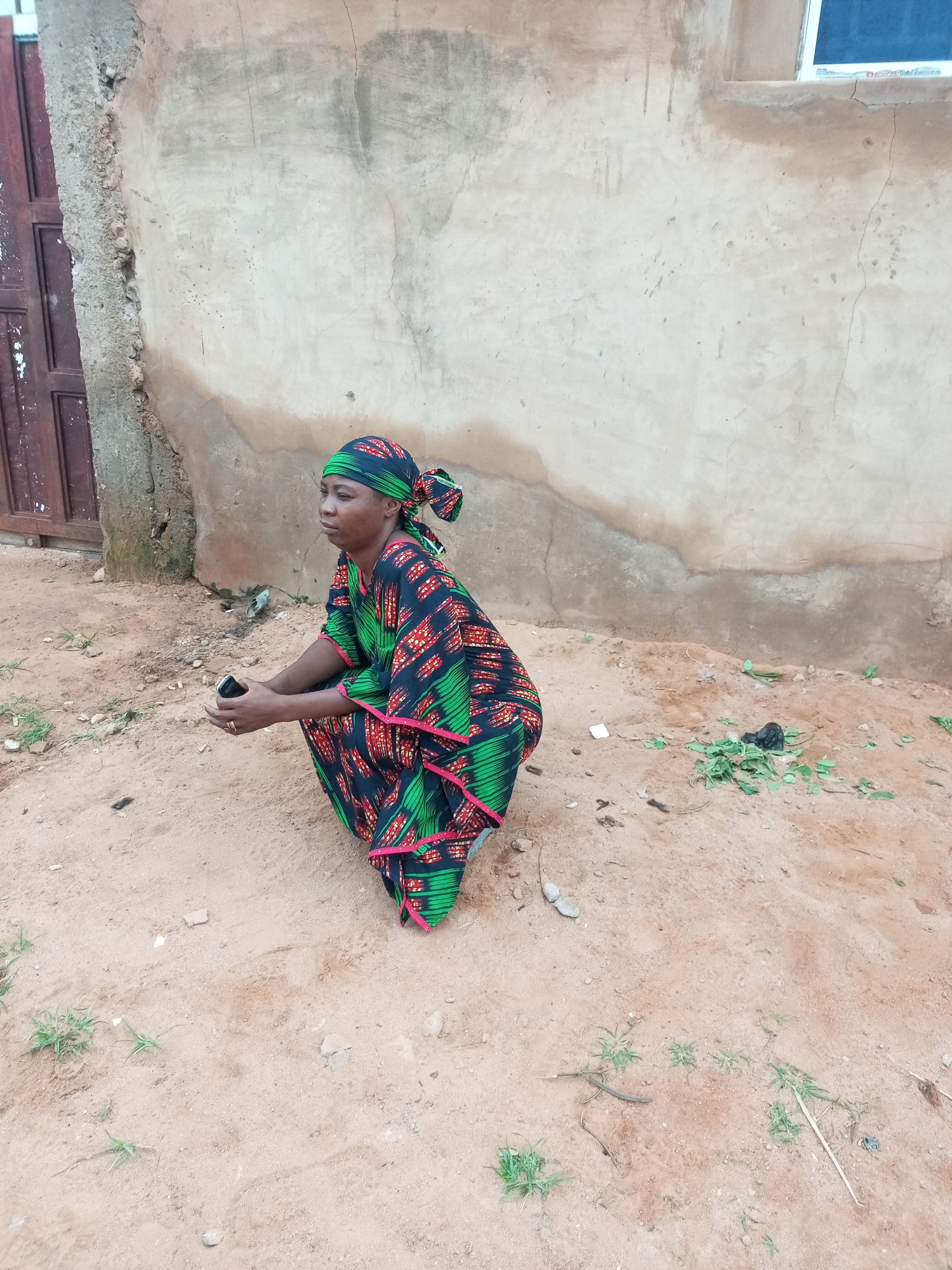 In Tempure, Nigeria, a woman kneels on sandy ground in front of a textured, light-colored wall. She is wearing a dark blue headscarf with a geometric pattern in green and red, matching the design of her long, flowing dress. The dress also features pink trim. Her hands are clasped, and she is holding a dark rectangular object, possibly a mobile phone. The wall behind her shows signs of age and weathering, with some cracks and staining. To her left, a portion of a brown door or gate is visible. The ground is mostly sand with sparse green vegetation and a few small stones. A small black plastic bag and some green leaves are scattered on the sand to her right.