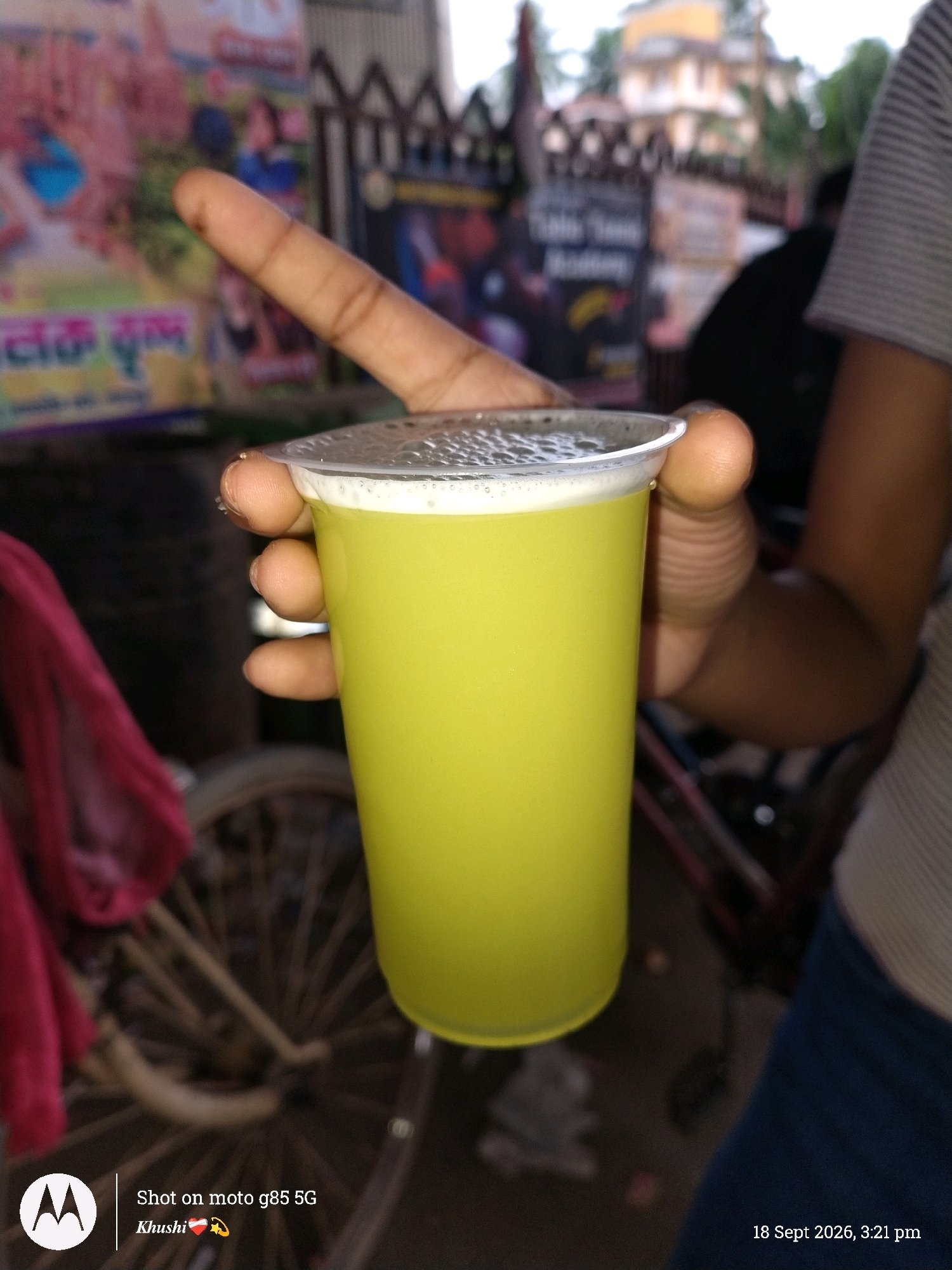 The image shows a close-up of a hand holding a clear plastic cup filled with a light green, frothy liquid, likely sugarcane juice. The cup is partially covered by the fingers of a person's hand. In the background, there is a blurred scene suggesting an outdoor market or street environment in Barrackpore, India. Visible elements in the background include posters with text in a non-English script, a bicycle wheel, a red cloth draped over something, and buildings. The bottom left of the image shows a "Shot on moto g85 5G" watermark with "Khushi" and two emojis. The bottom right corner displays the date and time: "18 Sept 2026, 3:21 pm". The overall impression is a candid shot capturing a moment of refreshment in a local Indian setting.