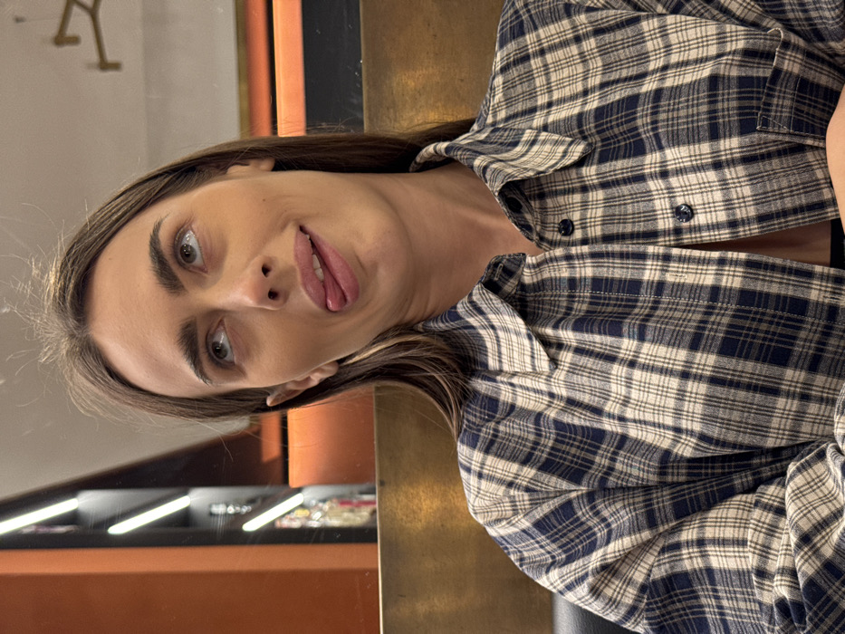 A single female individual is depicted from the chest up in an indoor establishment in Verona, Italy. She has medium-long, dark brown hair, fair skin, and prominent eyebrows. Her eyes are rolled upward and to the right, while her tongue is extended from her mouth and angled slightly to the left, indicating a playful expression. She is wearing a blue and beige plaid long-sleeved button-down shirt, unbuttoned at the top to reveal a dark garment underneath. The background consists of a horizontal golden-toned surface directly behind her. To the upper left, a dark vertical panel is visible, featuring two illuminated white linear light fixtures. To the upper right, a reflective surface shows a blurred, golden-colored, angular object, possibly a fixture or decorative element within the establishment.