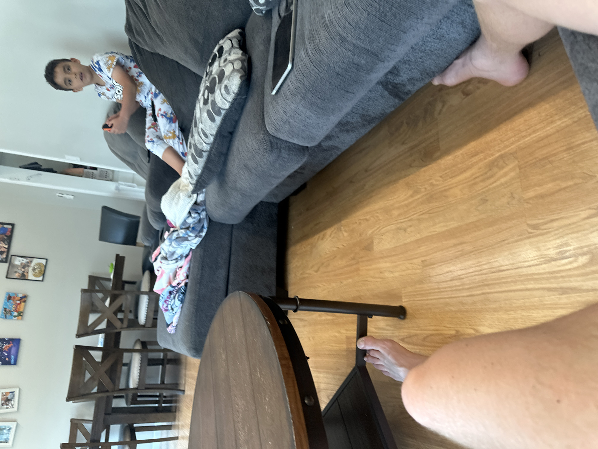 A room interior in Pepper Pike, United States of America, shows a boy in patterned pajamas holding a controller on a gray sofa. A round, dark wood table with a metal frame is present. A partial view of a person's legs and feet appears in the foreground and background. Chairs are visible in the background. A wall with framed images is on the left.