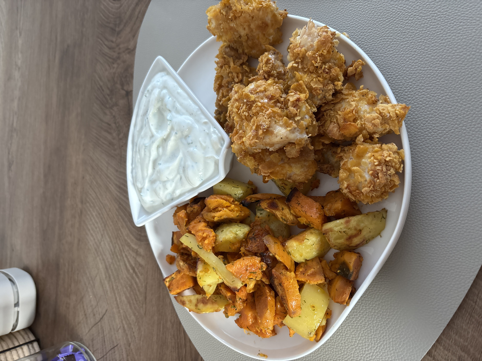 A close-up, high-angle shot captures a plate of food on a wooden table in Viernheim, Germany. The white, oval plate is divided into three sections, holding roasted vegetables, crispy chicken pieces, and a small dish of dipping sauce. The vegetables, appearing to be sweet potatoes and potatoes, are cut into irregular chunks and seasoned with herbs. They are arranged on the left side of the plate. To the right, golden-brown chicken tenders, coated in a crunchy breading that resembles crushed cereal or cornflakes, are piled high. A triangular white dish filled with a creamy, herb-flecked sauce sits at the top of the plate, between the vegetables and chicken. The plate rests on a light gray placemat, which is on a wooden table with a visible grain pattern. In the background, a portion of a white kitchen appliance and the edge of a glass with purple objects are visible. There are no people or animals in the image.