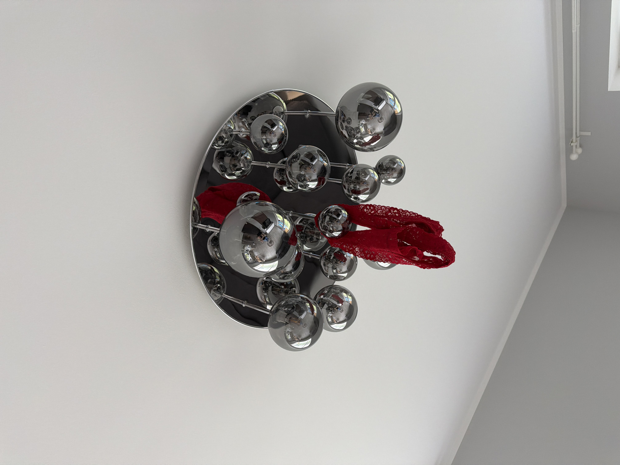 A wall-mounted decorative light fixture in the shape of a half-disc with chrome spheres attached by rods to the disc is adorned with a red lace garment. The fixture is mounted on a white wall, possibly in an interior space. The environment is a room with white walls and a corner where the wall meets the ceiling is visible. The image does not provide information about the location in Dreiliņi, Latvia.