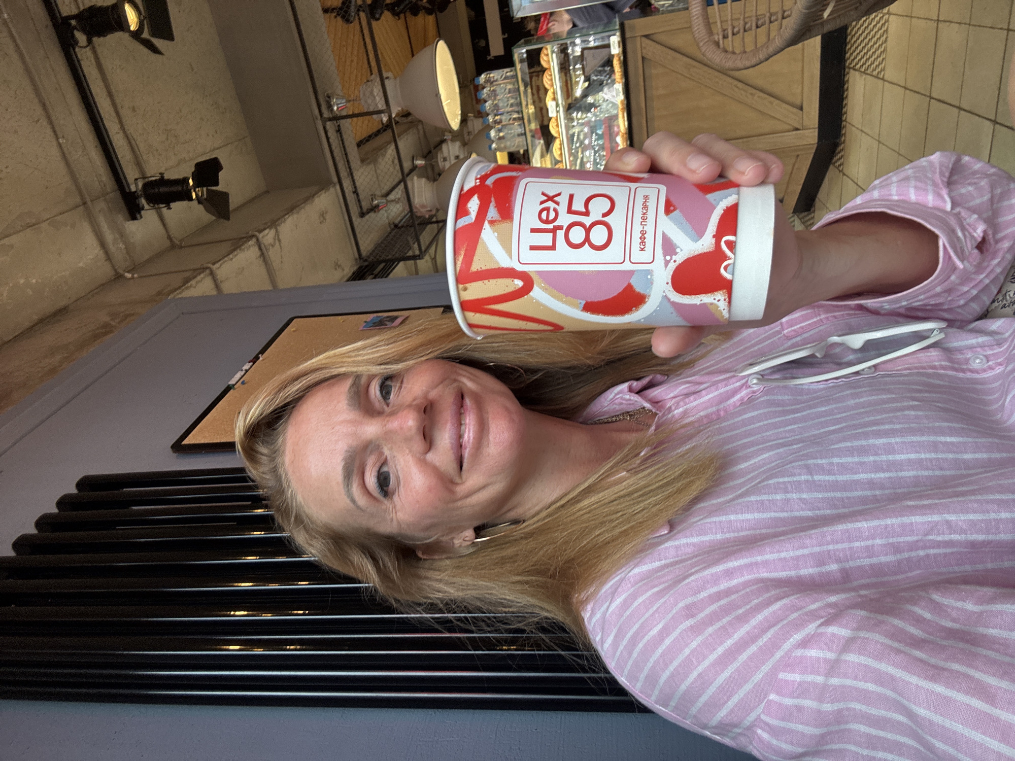 A woman with blonde hair and light skin is holding a paper coffee cup with a colorful design and Russian text that reads "Цех 85 кафе-пекарня." She is wearing a pink and white striped shirt and a necklace. The woman is smiling and looking at the camera. She appears to be in a cafe or bakery, with a display case of pastries in the background and industrial-style lighting overhead. The wall behind her has vertical black decorative elements. The scene suggests a casual outing in Saint Petersburg, Russia.