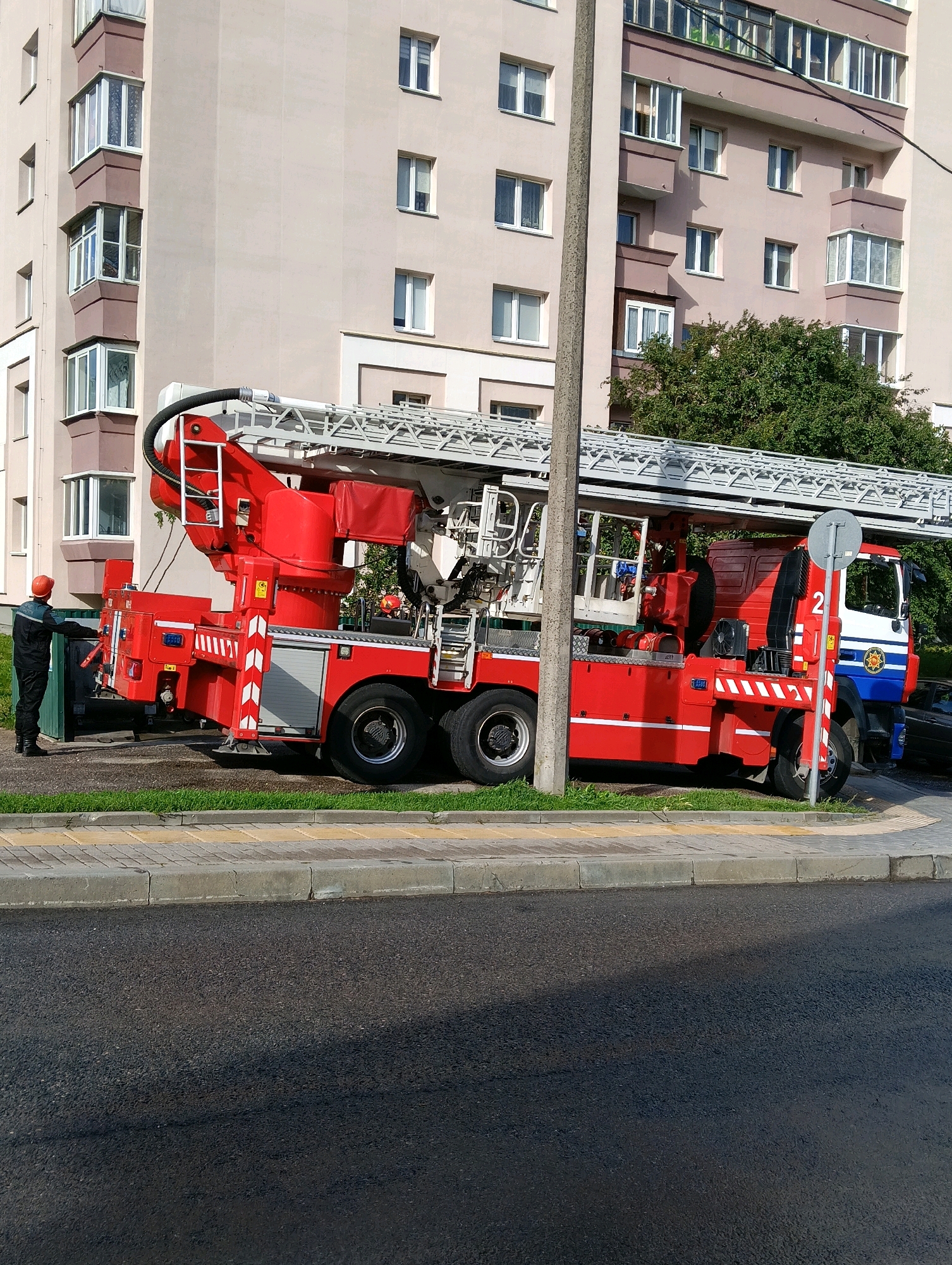 A red aerial ladder fire truck is parked on a paved road in front of a light-colored, multi-story apartment building in Hrodna, Belarus. A firefighter in a dark uniform and orange helmet is standing next to the rear of the truck, facing a green dumpster. The fire truck has a long, silver ladder extended above it. The apartment building has numerous windows and balconies. Trees are visible behind the fire truck. A light pole stands to the right of the truck. The ground in front of the building is a sidewalk with yellow and gray paving stones, bordered by a curb. The road surface is asphalt.