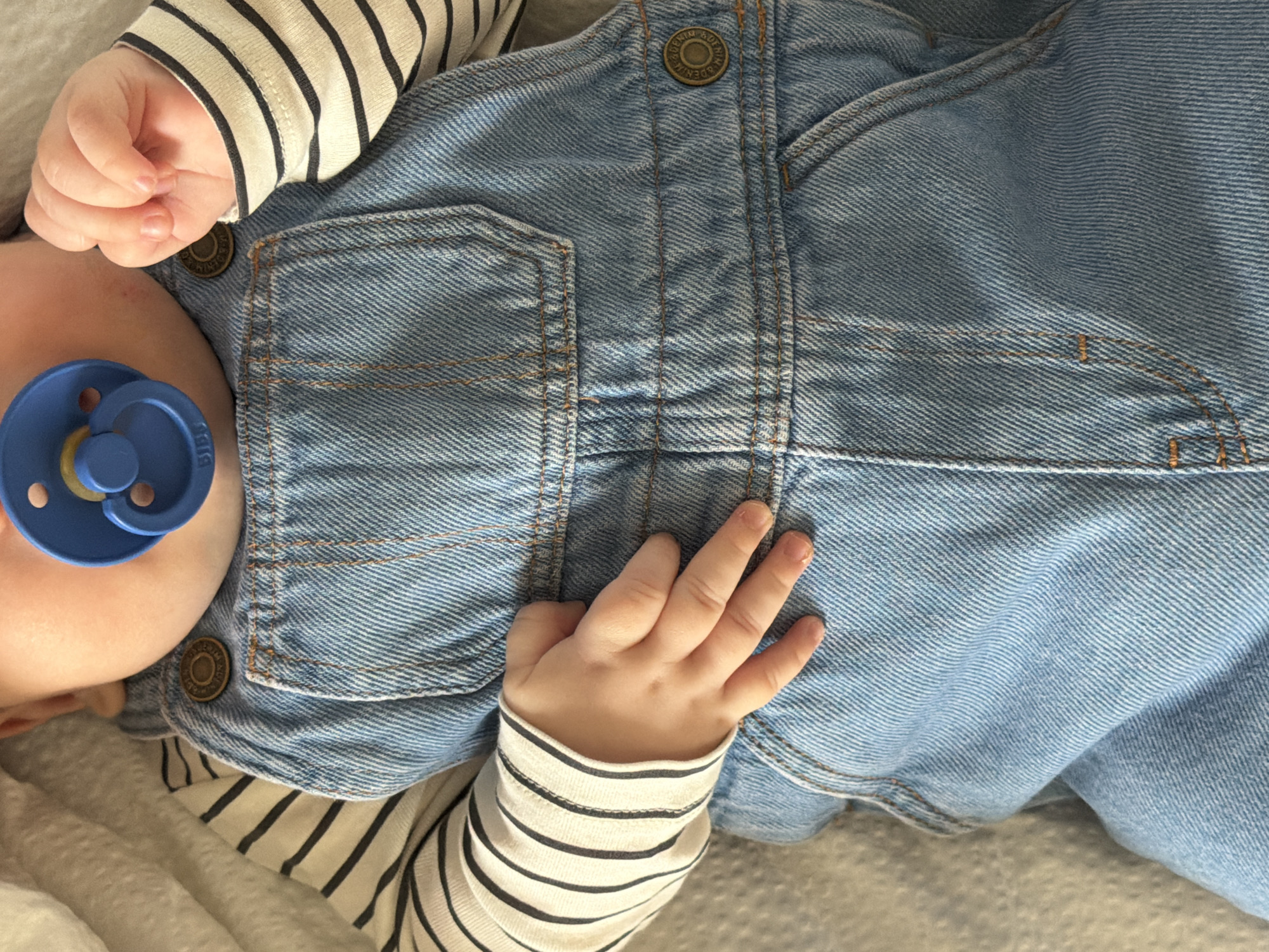 A close-up, high-angle shot depicts a baby lying down, partially visible from the chest upwards. The baby wears a long-sleeved shirt with alternating off-white and black horizontal stripes, covered by light blue denim overalls. A blue pacifier with a yellow nipple is in the baby's mouth, with the brand name "BIBS" visible on its front. One of the baby's hands, with fingers gently curled, is positioned near the pacifier, while the other rests on the denim fabric.
The denim overalls feature brass-colored buttons, some clearly engraved with the text "PREMIUM DENIM." Detailed stitching in a contrasting lighter thread is visible on the pockets and seams of the denim. The baby is resting on a soft, light-colored, textured surface, likely a blanket or bedding, suggesting an indoor setting. The image evokes a sense of calm and comfort. While no specific time of day or weather is evident, the scene is situated in Khimki, Russia, as per the provided context.