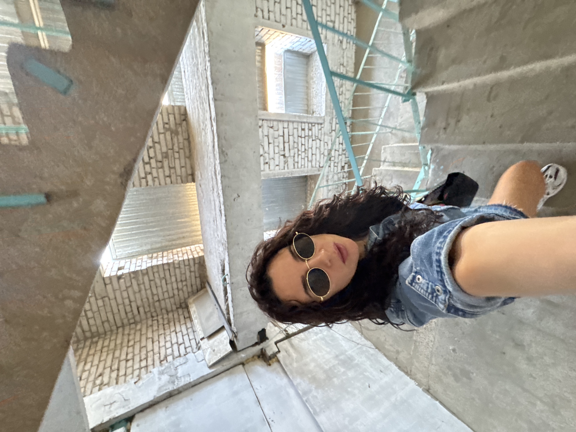 A woman wearing sunglasses and a denim jacket stands on concrete stairs within a stairwell, possibly in the city of Chaikovsky, Russia. The stairwell is unfinished, with brick walls and metal railings. The view is an overhead shot of the woman, with the stairwell and the brick construction forming the background. The woman is holding the camera at arm's length, capturing a selfie.
