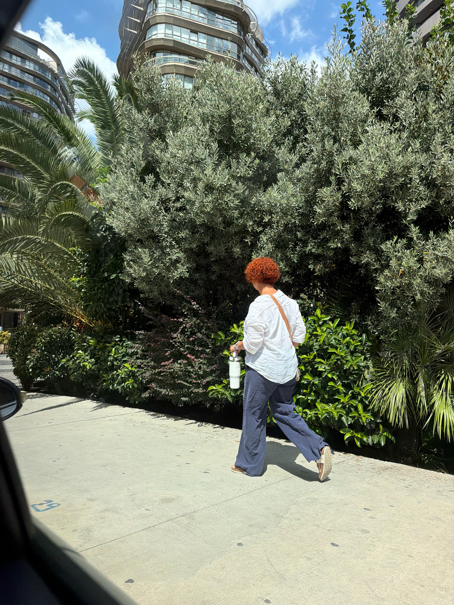 An individual with bright red, curly hair, wearing a white linen shirt and dark blue wide-leg pants, walks along a concrete sidewalk. They are carrying a white water bottle in their left hand and have a light brown crossbody bag slung over their right shoulder. The person is walking away from the viewer, towards the right side of the frame. The sidewalk is bordered by lush green foliage, including palm trees and a dense, leafy shrubbery. In the background, two modern, curved-facade buildings rise against a blue sky with scattered white clouds. The scene appears to be in an urban environment, possibly Bakırköy, Turkey, given the presence of palm trees and contemporary architecture. The lighting suggests it is daytime.